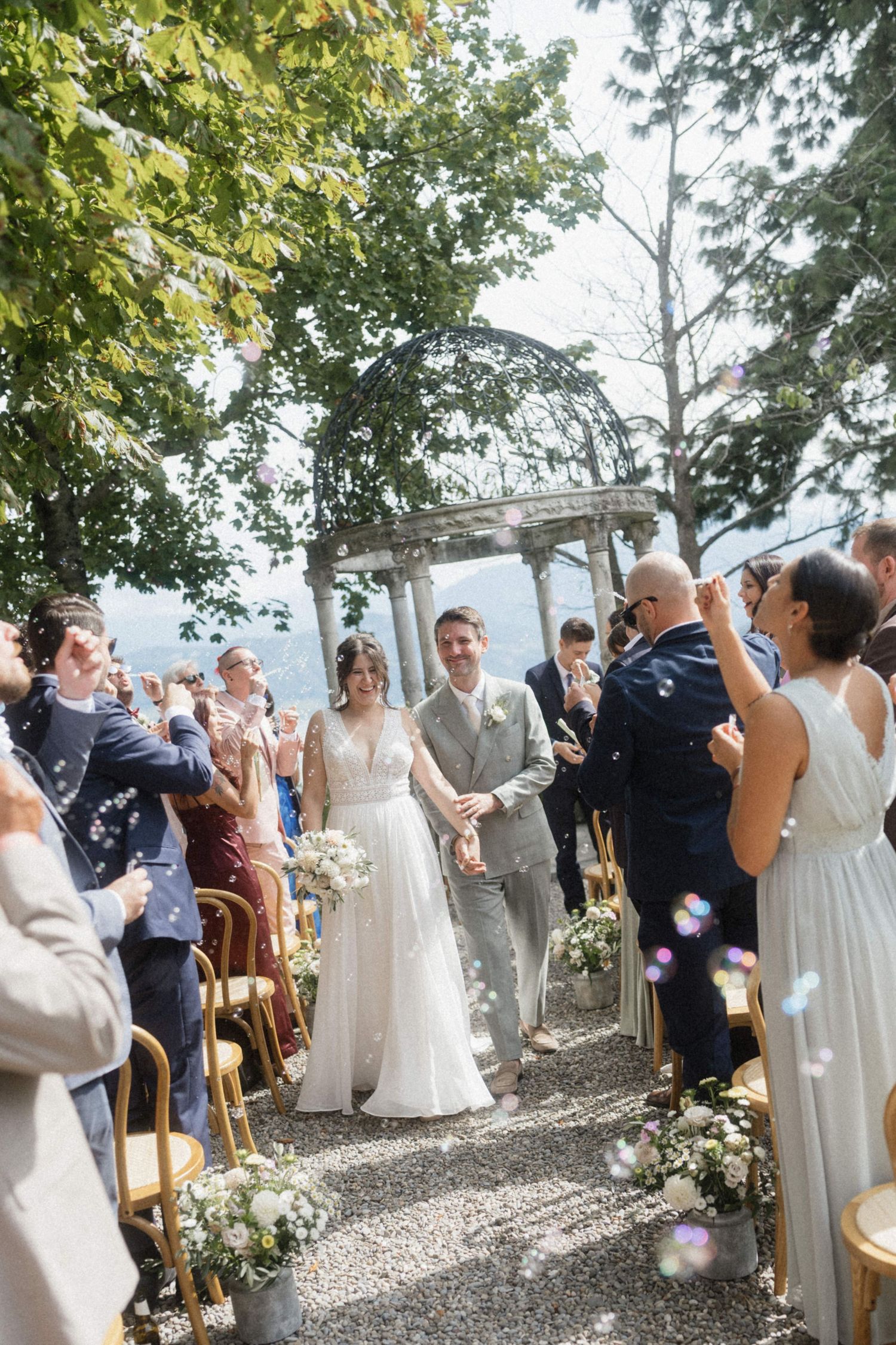 Newlyweds walk down aisle through bubbles and cheering guests at outdoor ceremony.
