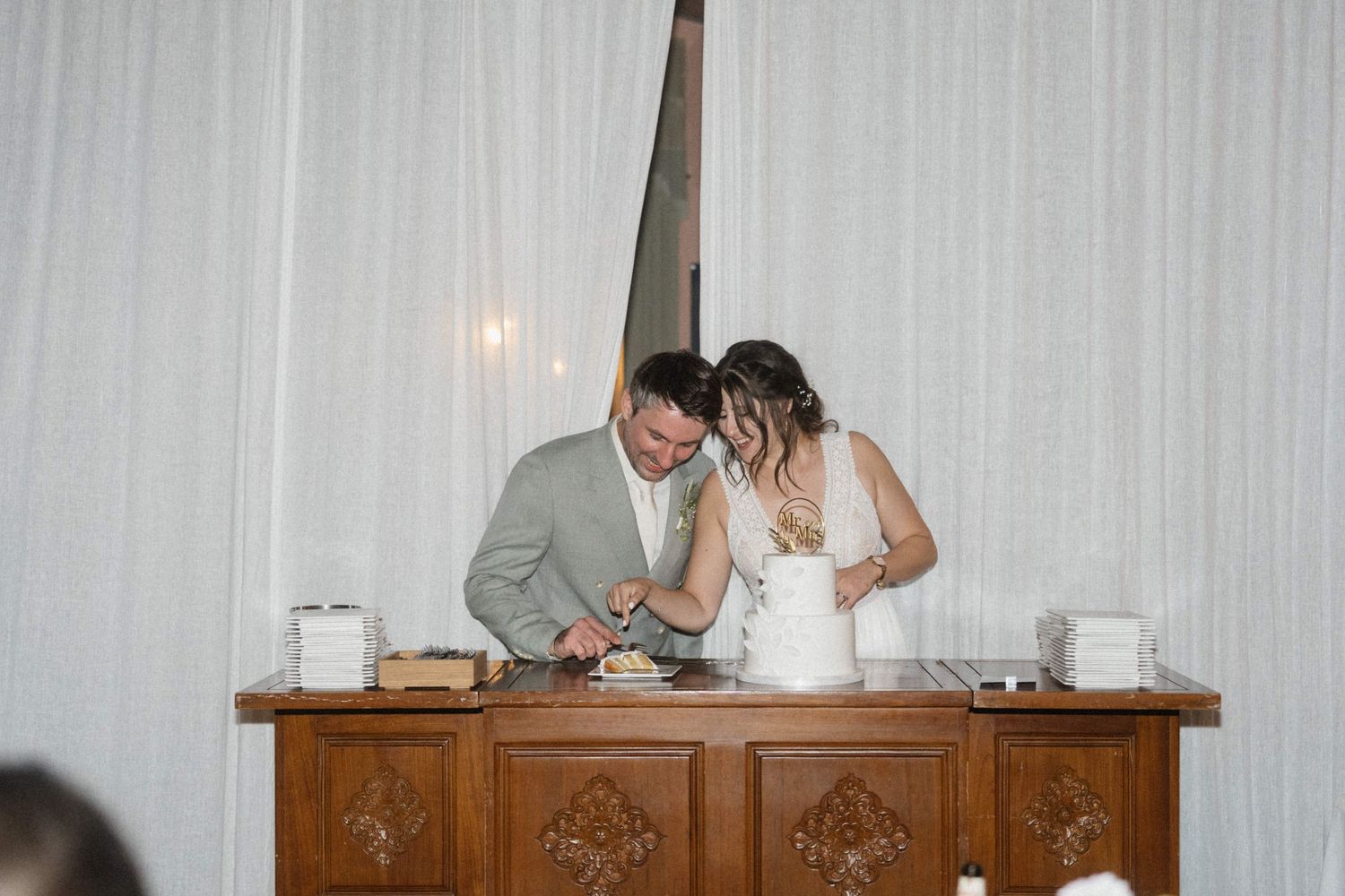 Bride and groom cutting their wedding cake together at a wooden table with white drape backdrop.