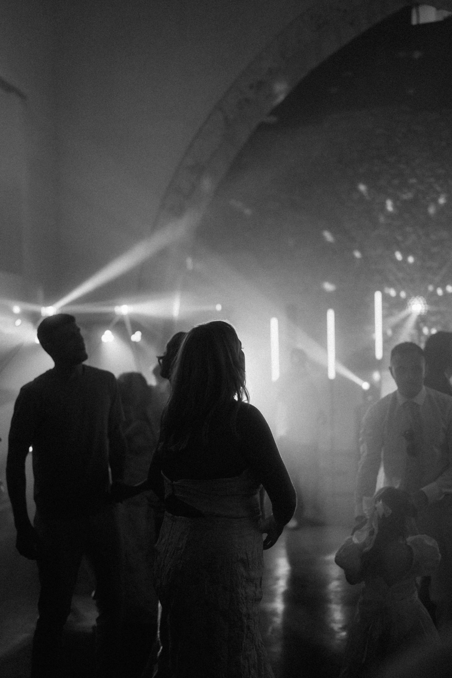 Black and white photo of silhouetted guests dancing at a wedding reception with dramatic stage lighting.
