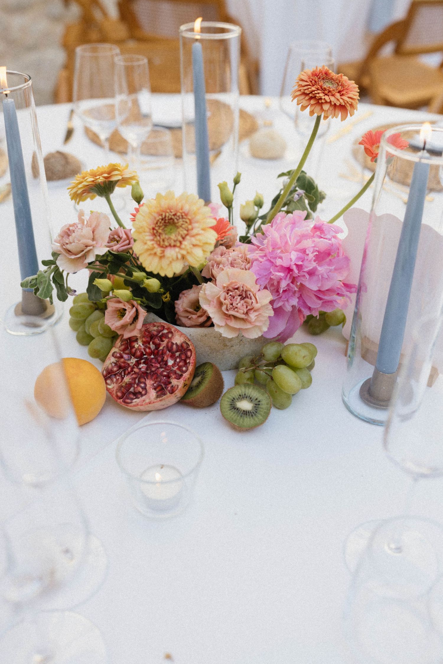 Elegant wedding table centerpiece with pink flowers, fruit, blue taper candles, and glassware on white linen.