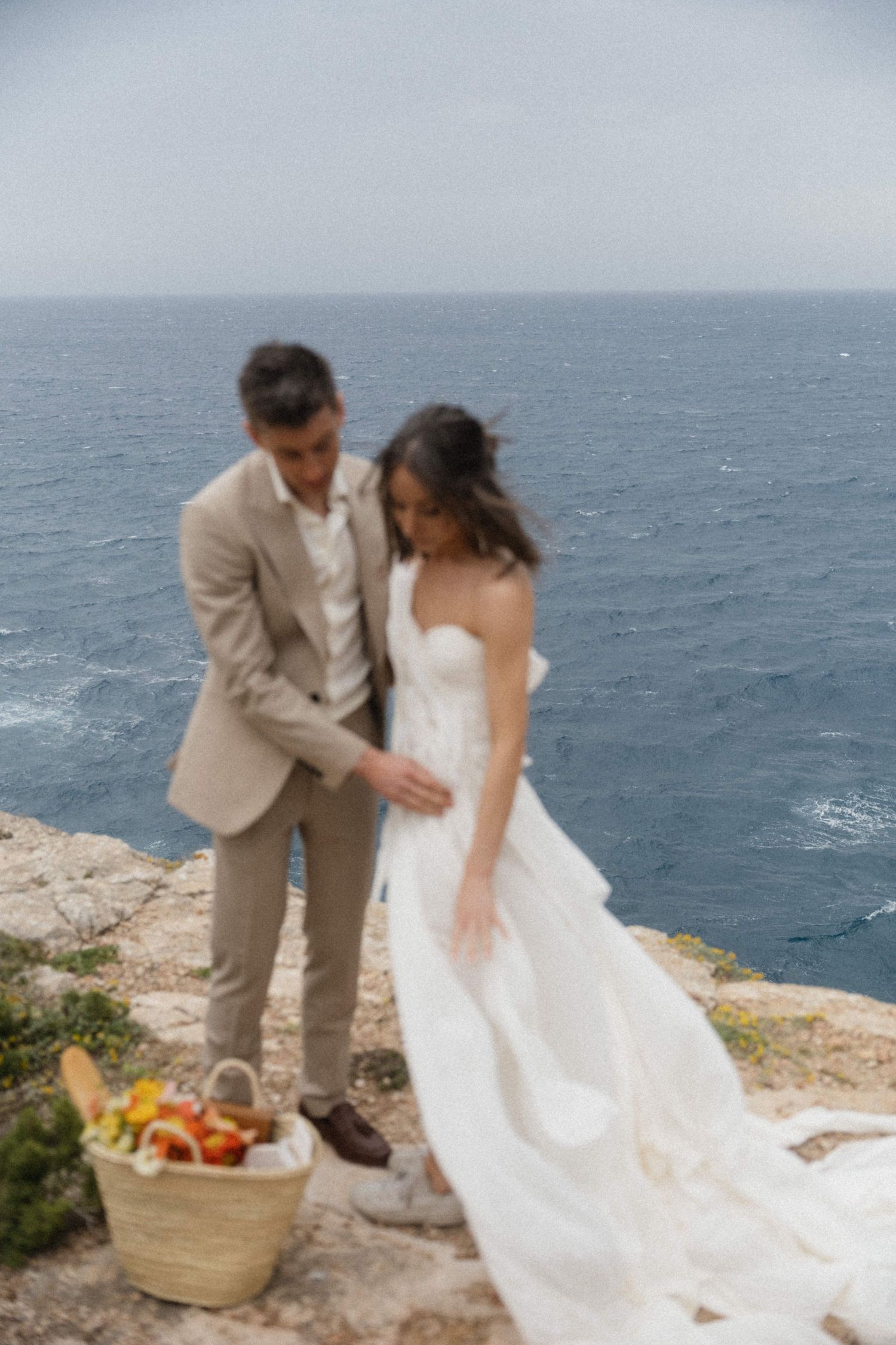 Bride and groom on ocean cliffside with picnic basket of fresh fruits and flowers nearby.