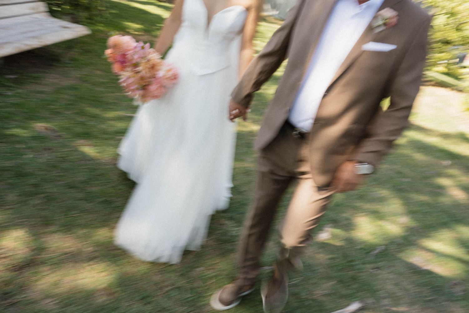 Blurred motion shot of a bride in white gown holding pink bouquet and groom in brown suit walking on green grass.