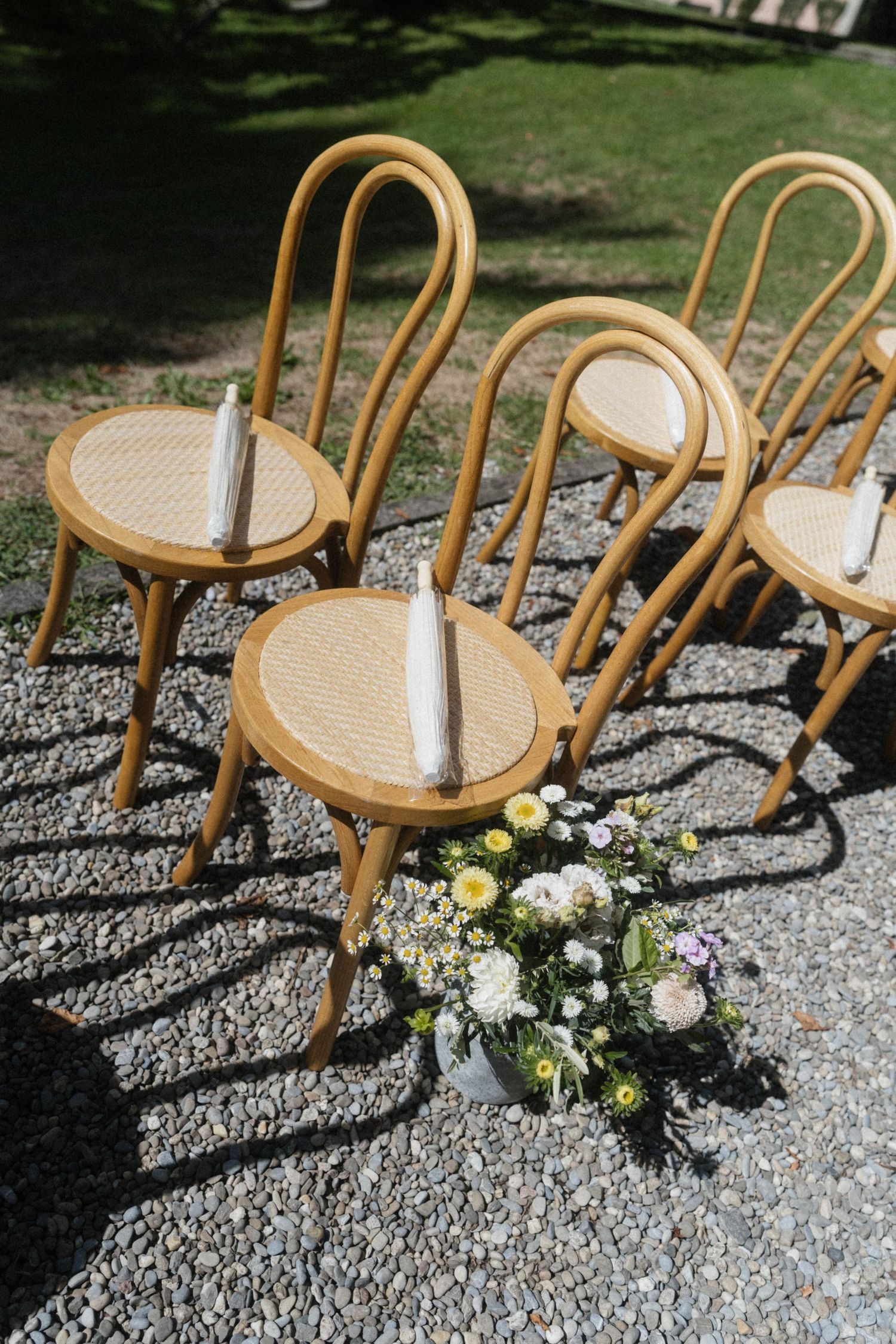 Elegant bentwood chairs with woven seats arranged on gravel for an outdoor wedding ceremony, adorned with candles and fresh flowers.