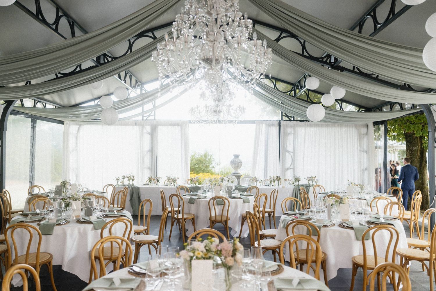 Elegant wedding reception tent with crystal chandelier, draped fabric ceiling, and round tables with gold chairs.