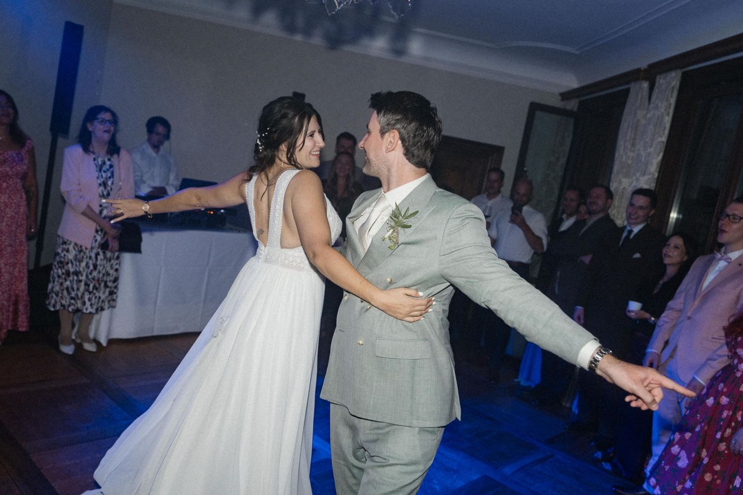 Bride and groom sharing their first dance under blue lights surrounded by smiling guests at their wedding reception.