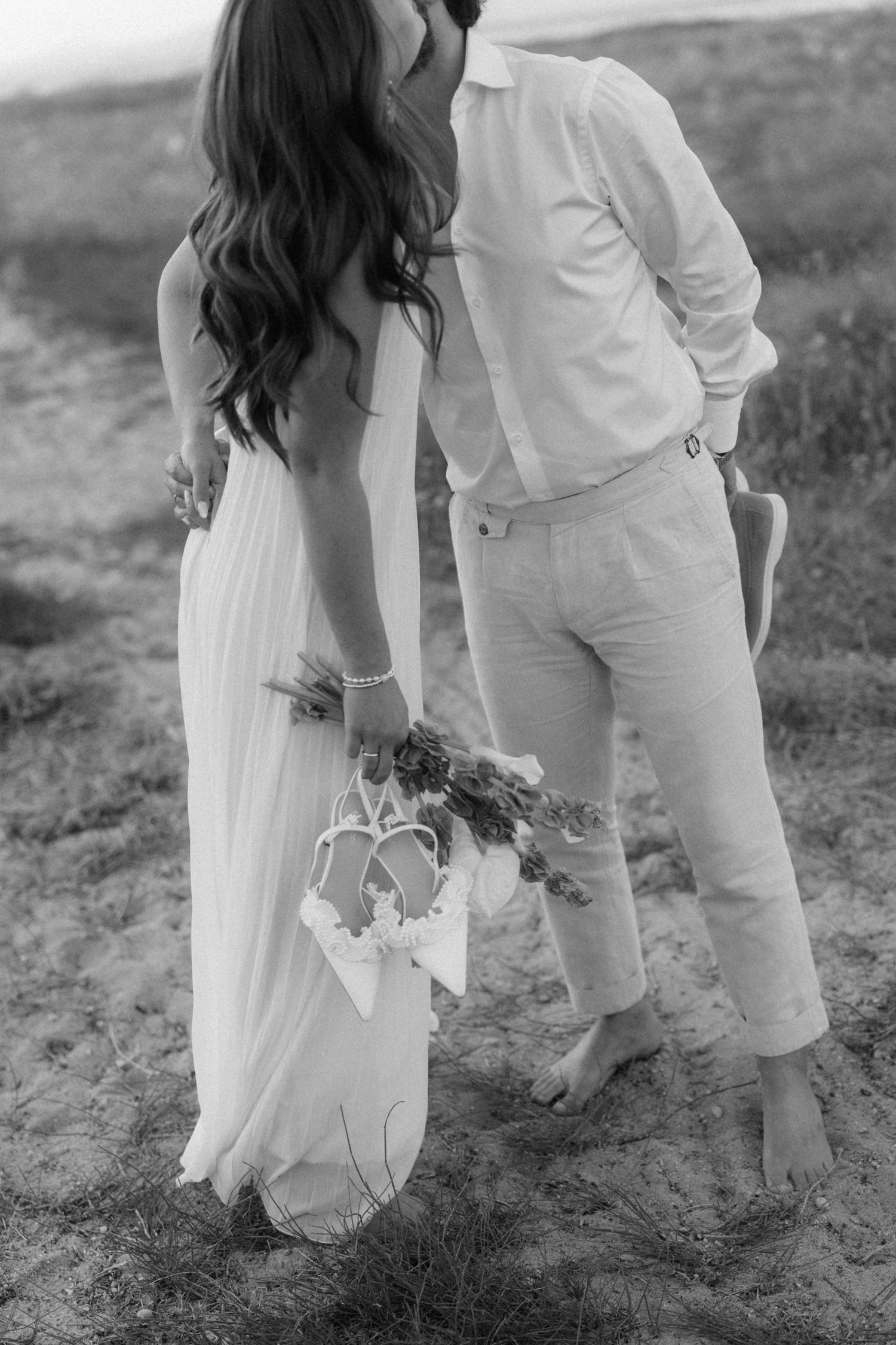 Black and white photo of couple embracing on sandy beach, bride holding bouquet and heels in backless dress.