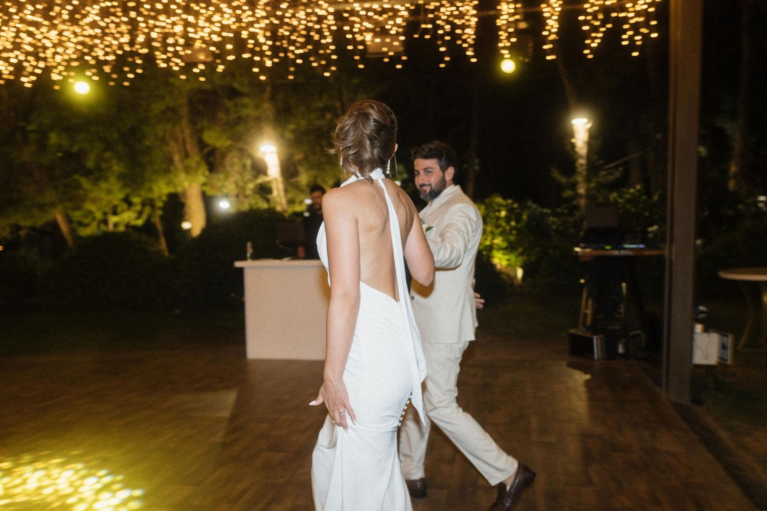 Bride and groom share first dance outdoors at night under warm string lights at elegant wedding reception.