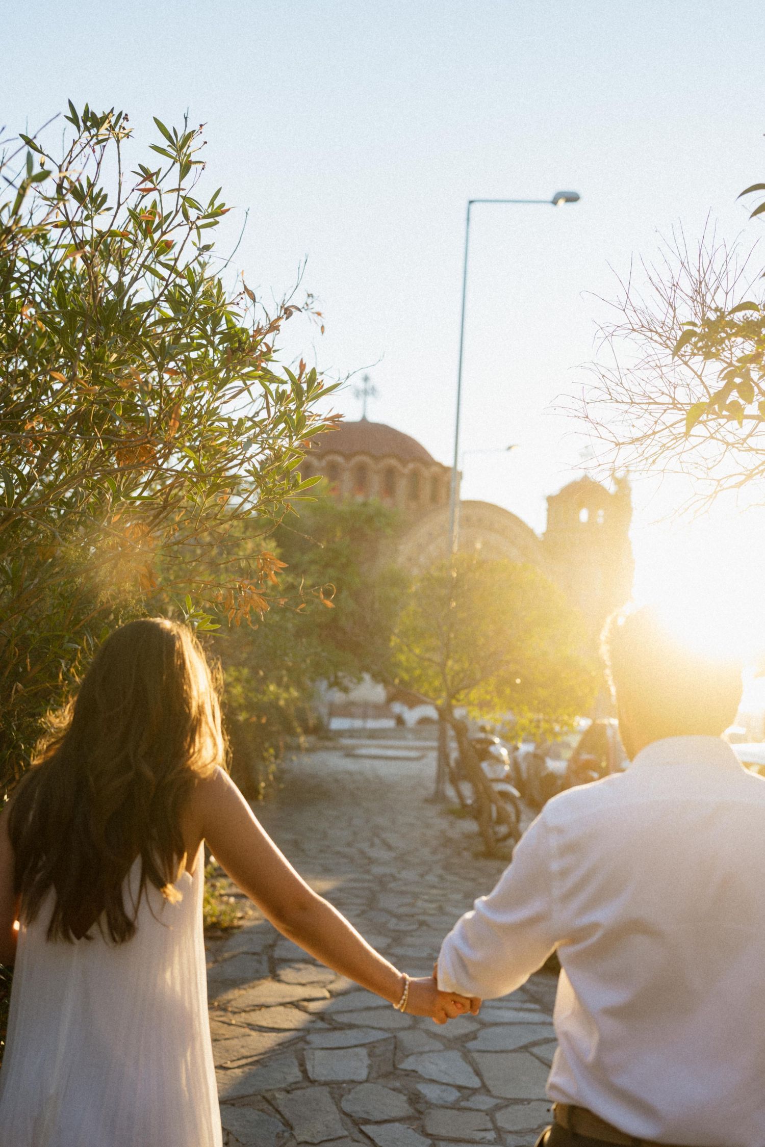 Couple holding hands walking toward a sunlit church at golden hour, surrounded by olive trees on a cobblestone path.