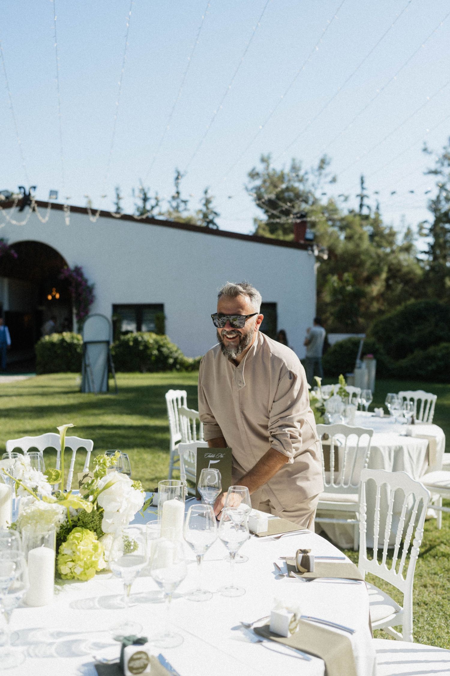 A smiling man in beige arranges glassware at an elegant outdoor wedding reception with white chairs and floral centerpieces.