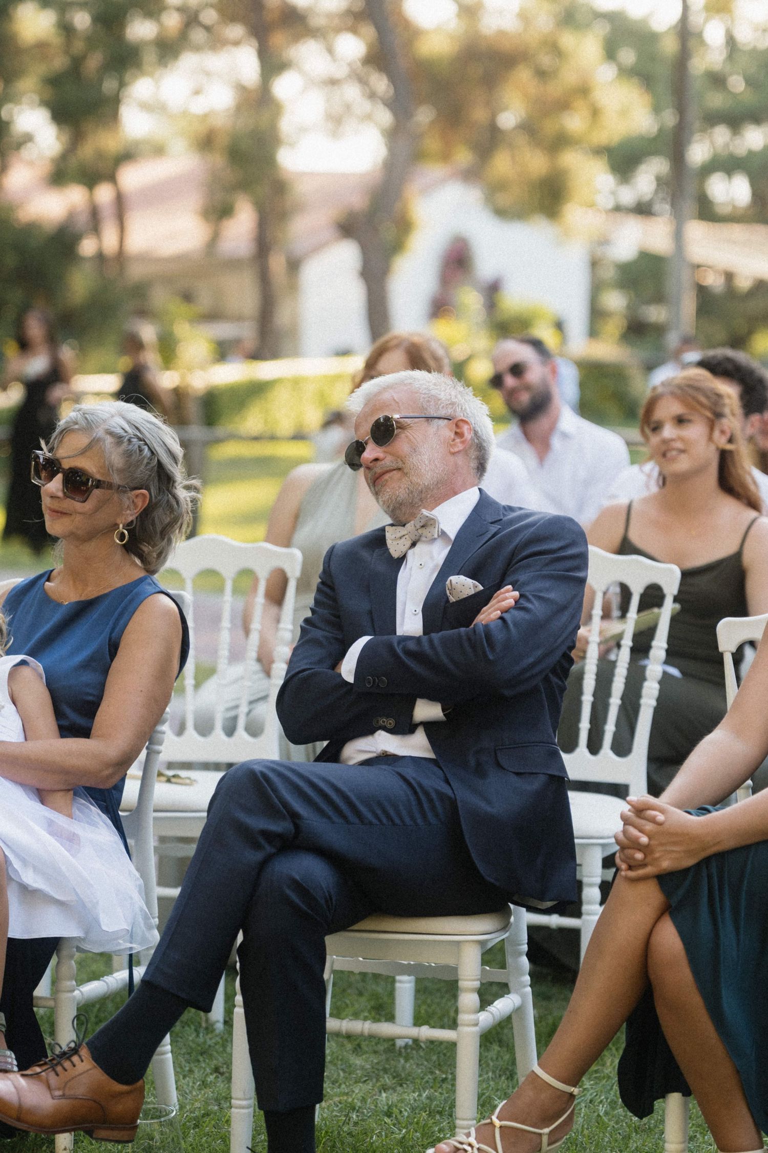 Elegant wedding guests seated outdoors, man in navy suit with bow tie and sunglasses sits with arms crossed.