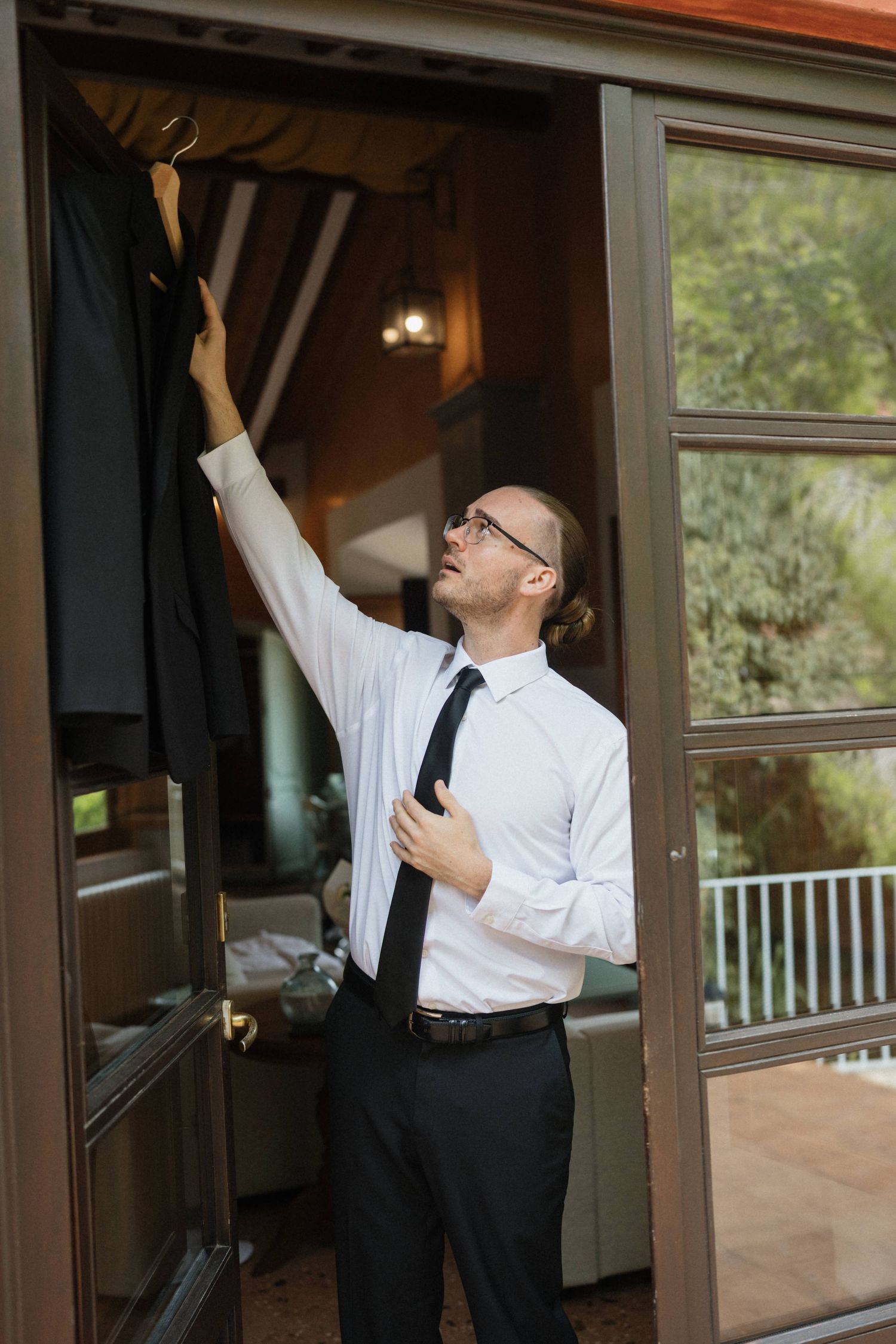Groom in white shirt, black tie and glasses reaches up to take his suit jacket from a glass door as he gets ready for the wedding.