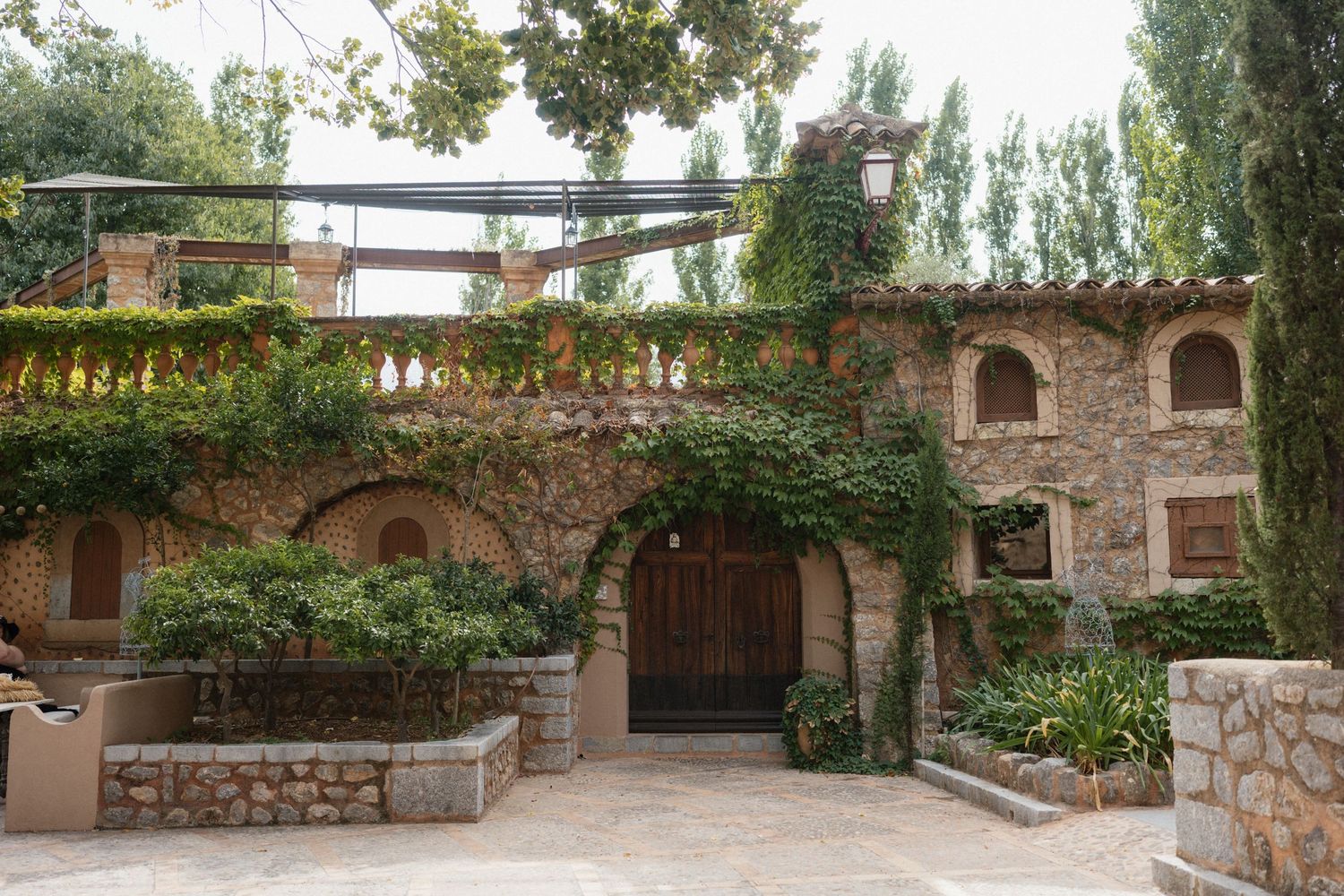 Rustic stone villa with a wooden arched door, ivy-covered walls and a sunlit terrace surrounded by lush green trees.
