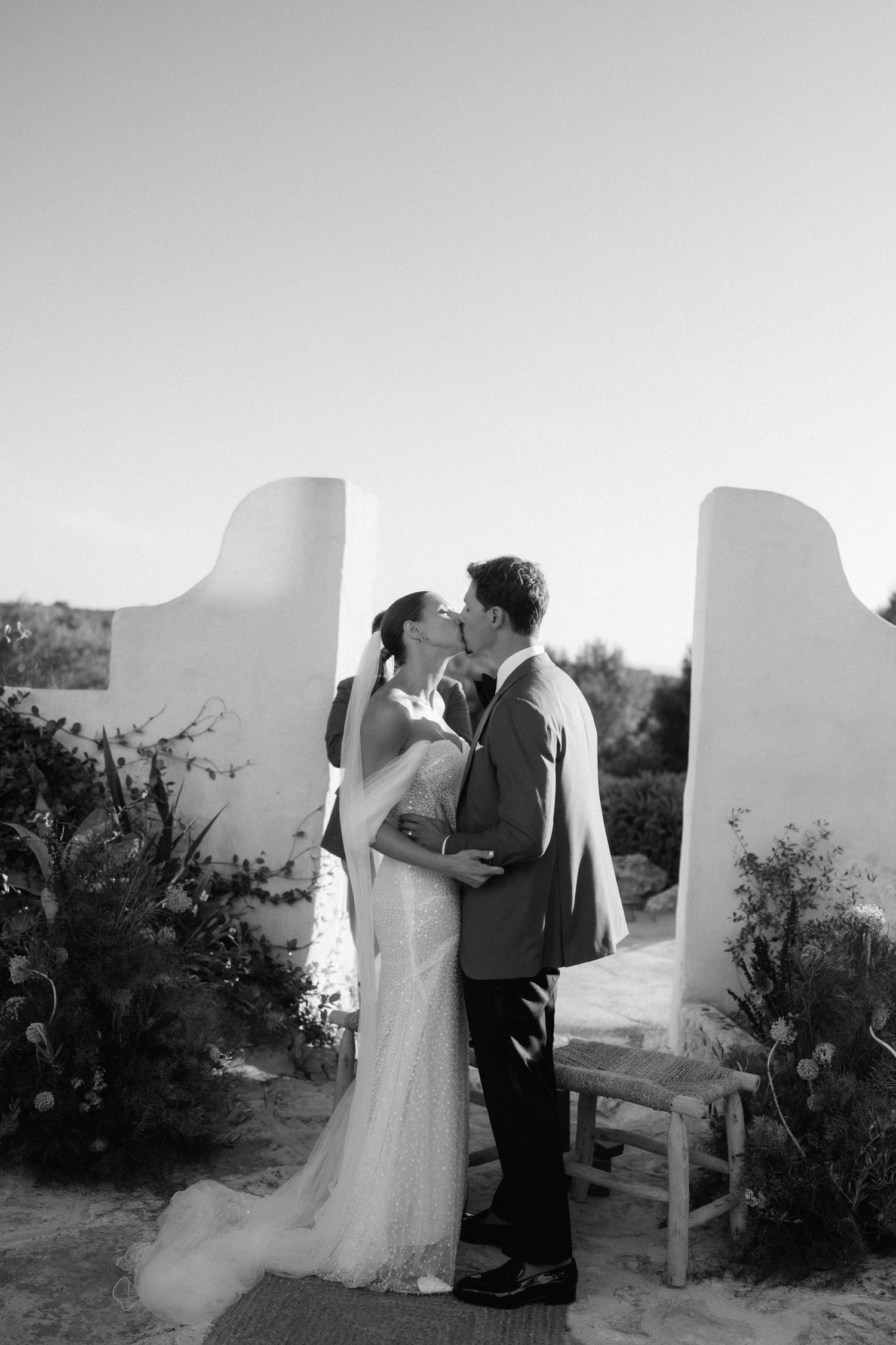 Black and white photo of a bride and groom kissing at an outdoor wedding ceremony between white stucco walls and flowers.