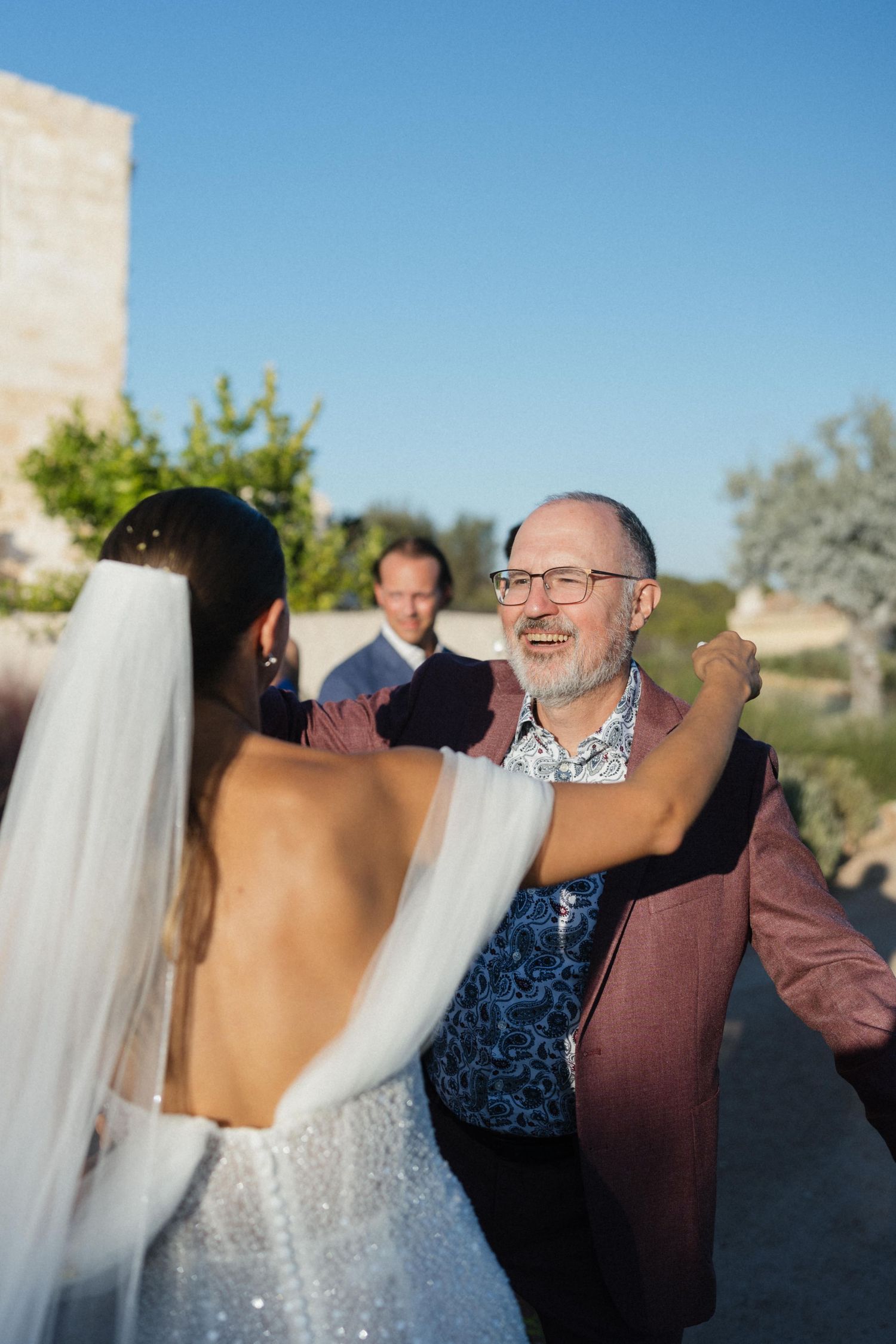 Bride in a sparkling backless gown and veil embraces a smiling bearded man in glasses and burgundy blazer outdoors.