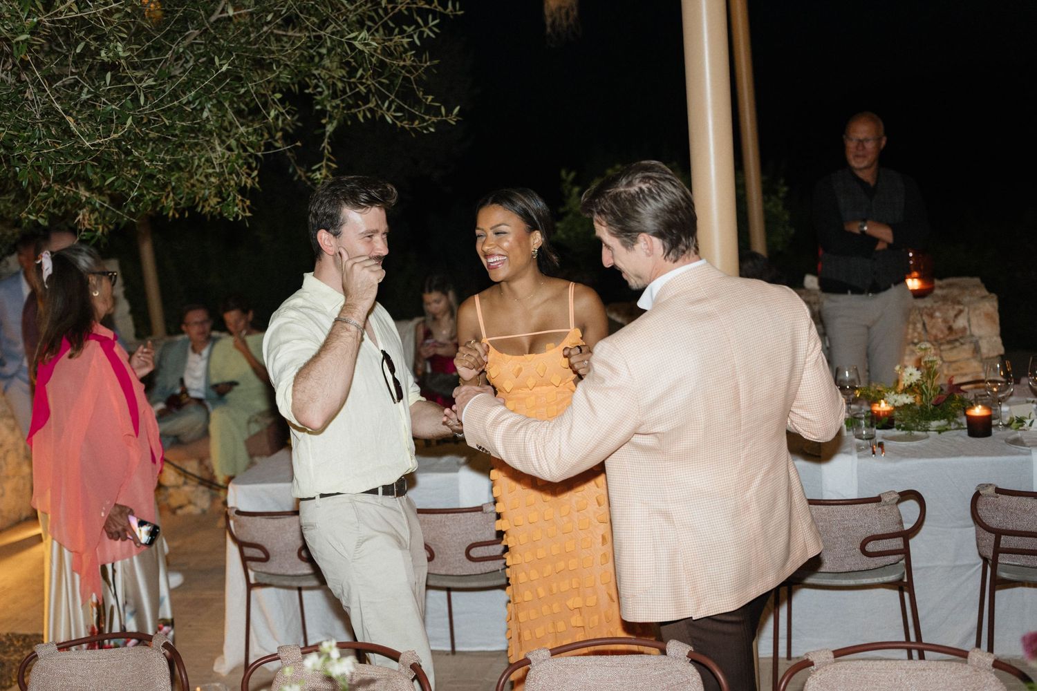 Wedding guests laugh and dance beside a candlelit dinner table at an outdoor evening reception.