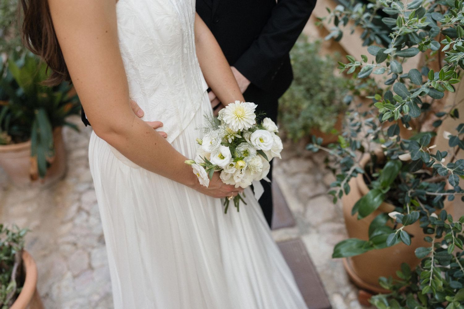 Bride in a white wedding dress holding a small bouquet of white flowers while embracing a groom in a dark suit near potted plants.