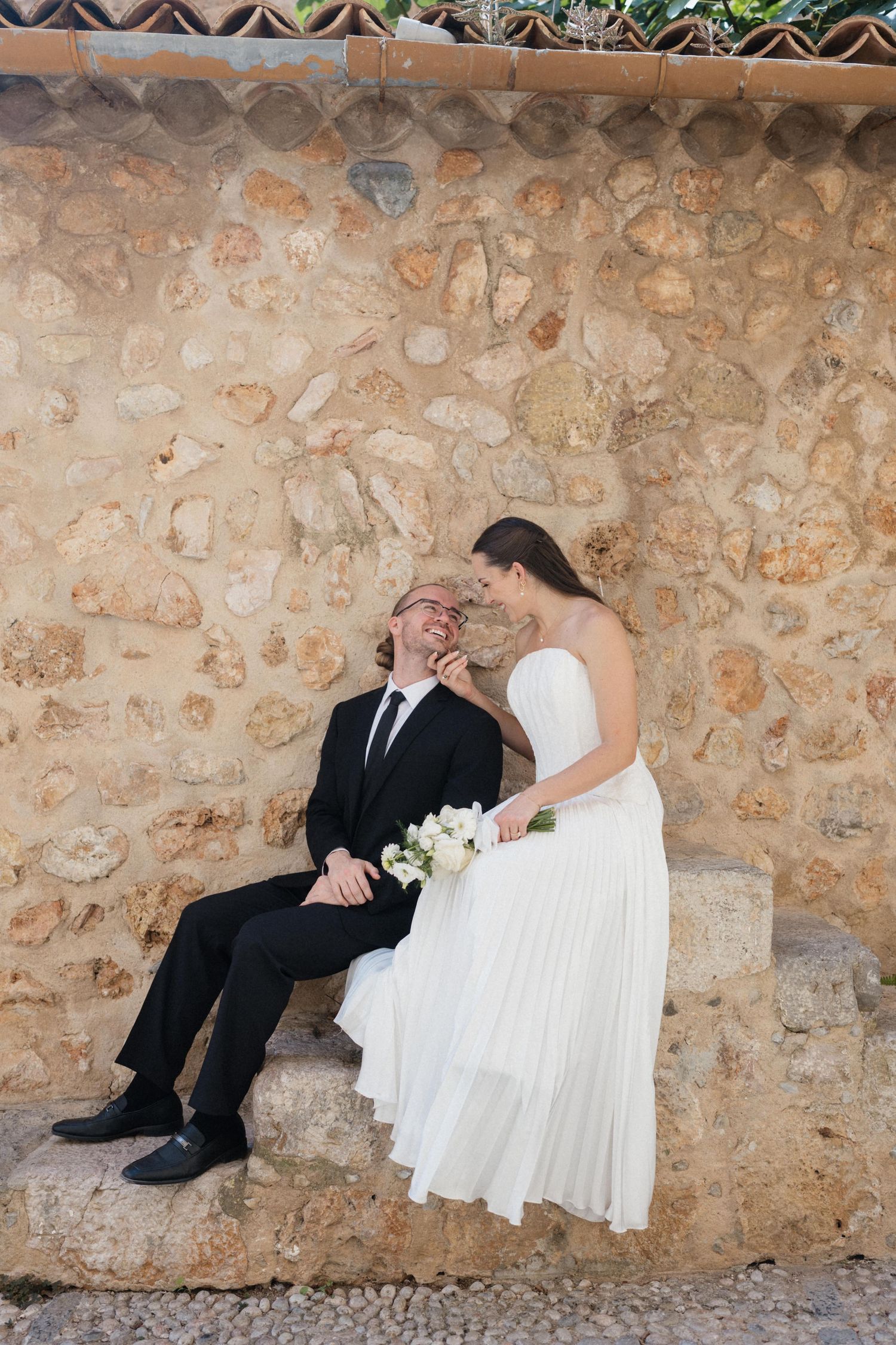 Bride in a white strapless gown holding a bouquet laughs with a groom in a dark suit while sitting on a stone ledge against a rustic wall.