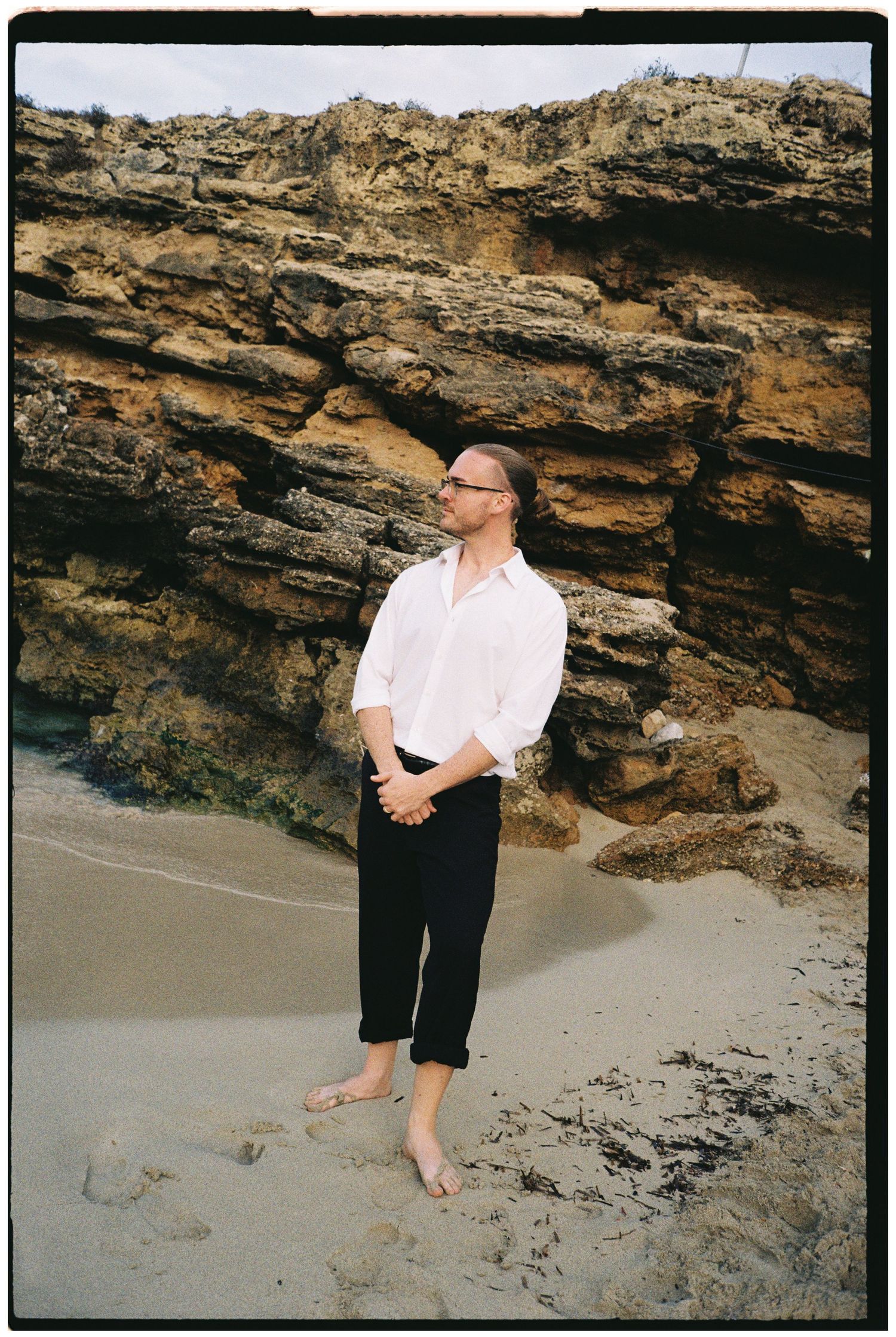 Barefoot man in a white shirt and black cropped pants stands on a sandy beach in front of layered golden rock cliffs.