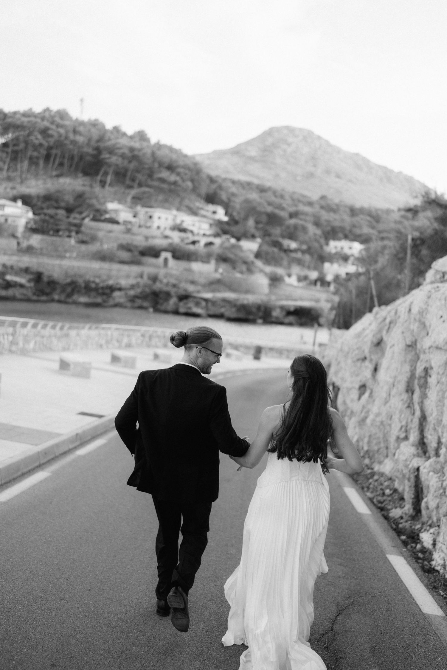 Black and white photo of a bride and groom holding hands and walking along a coastal road with hills and houses ahead.