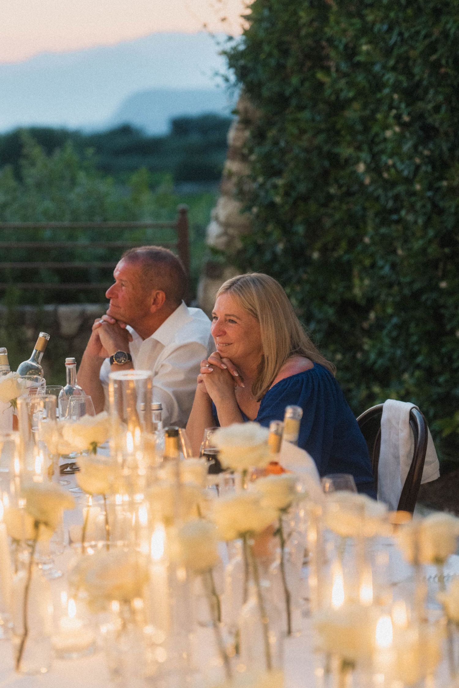 Couple seated at candlelit outdoor wedding reception table with white roses at dusk.