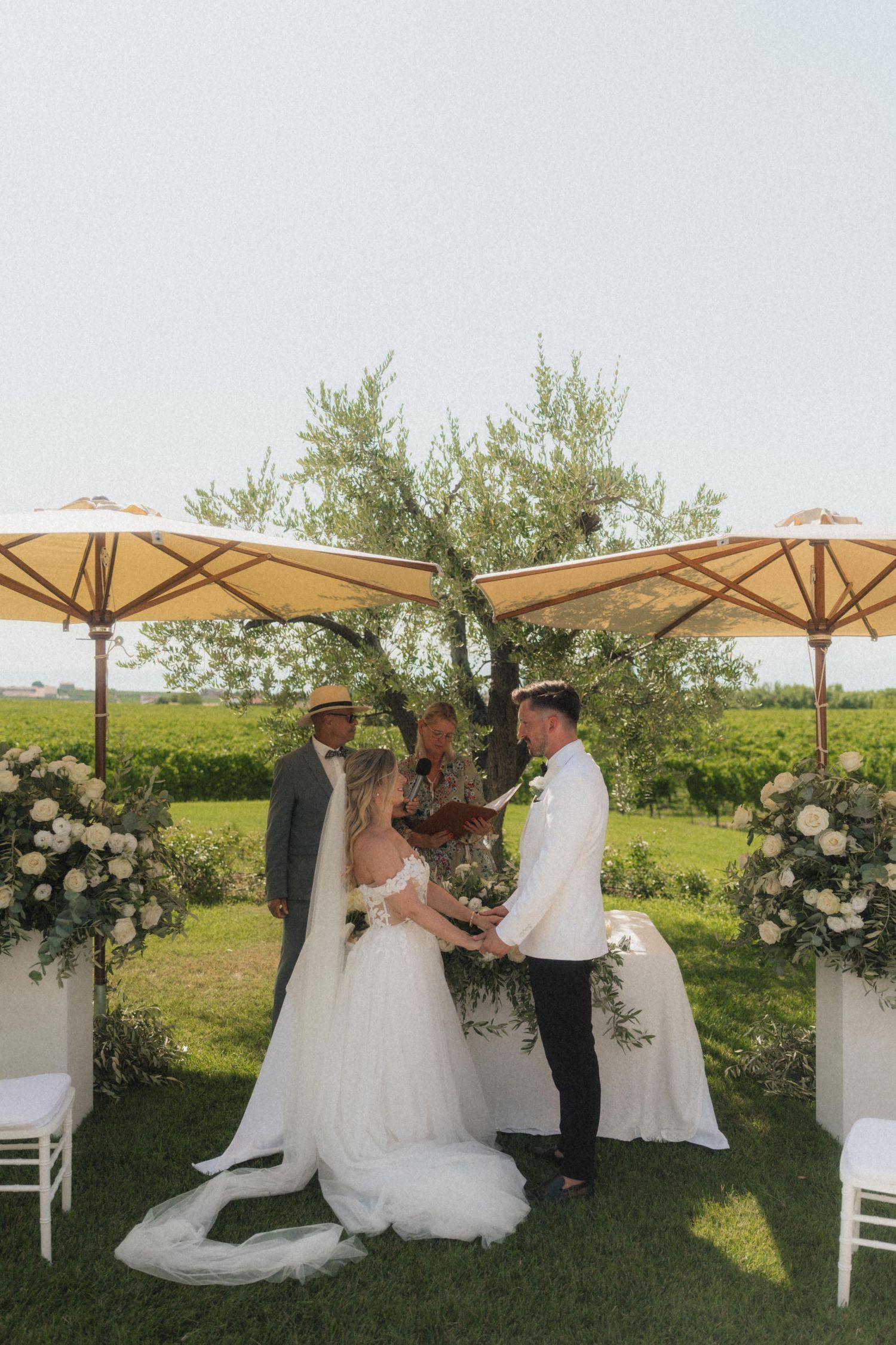 Bride and groom exchange vows outdoors at a vineyard wedding ceremony with white floral arrangements and yellow umbrellas.