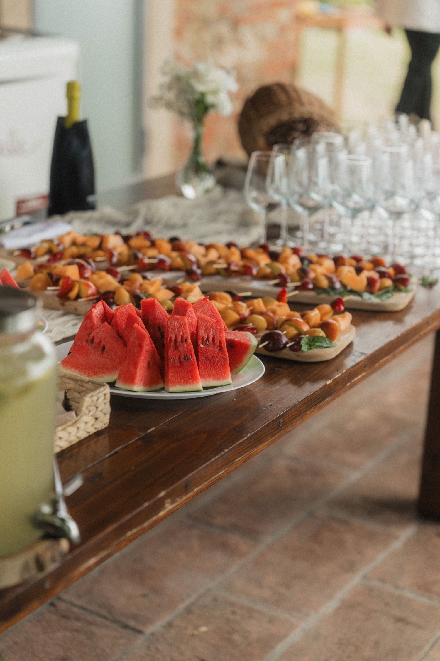 Elegant wedding reception buffet table with watermelon slices, fruit appetizers, wine glasses, and champagne bottle.