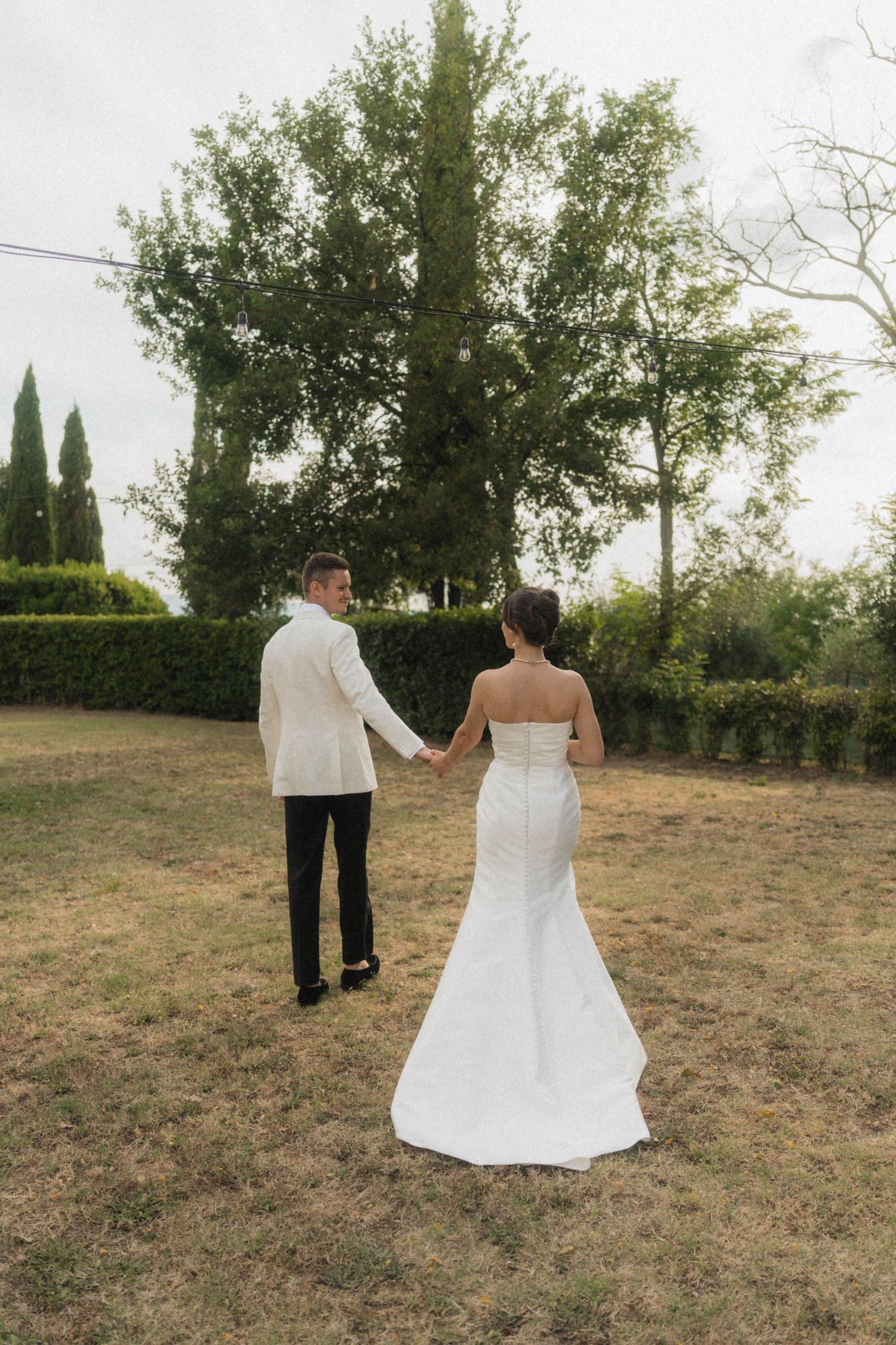 Bride and groom walk hand in hand through a lush green garden at their Tuscan wedding.