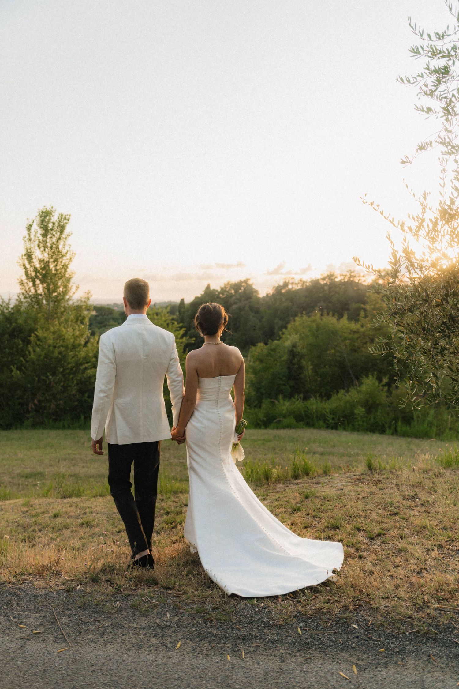 Bride and groom holding hands walking toward sunset in lush green countryside.
