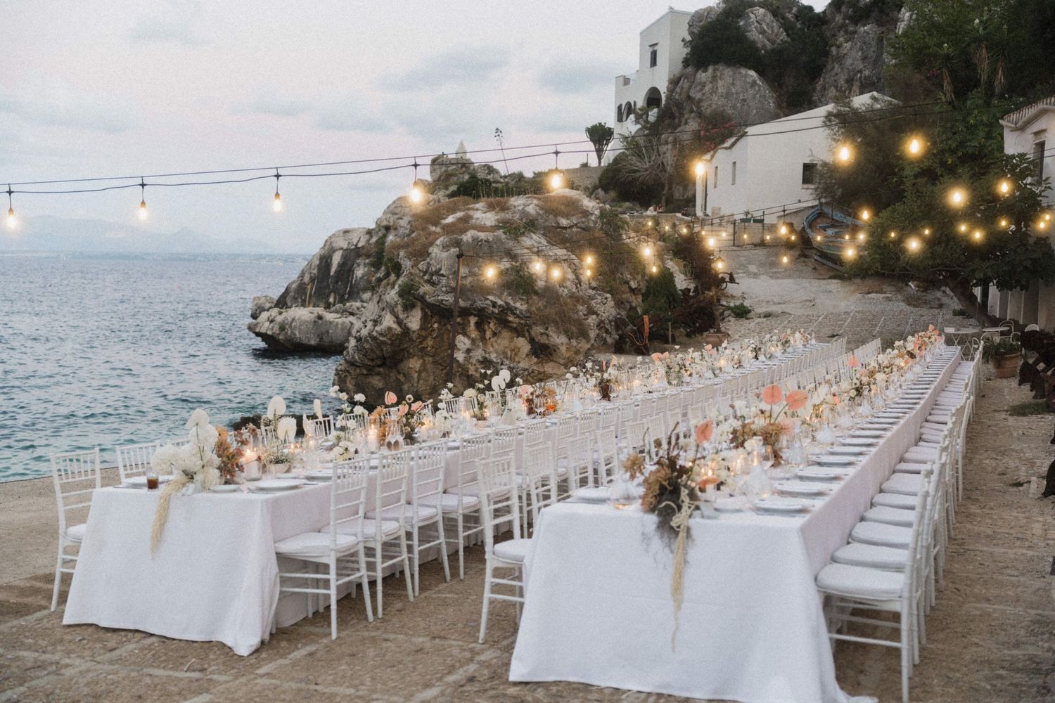 Elegant outdoor wedding reception with long white tables set by the sea at dusk.