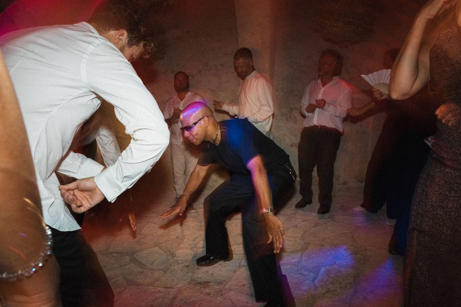 Energetic party guests dancing under blue and purple lights in a rustic stone-walled venue.