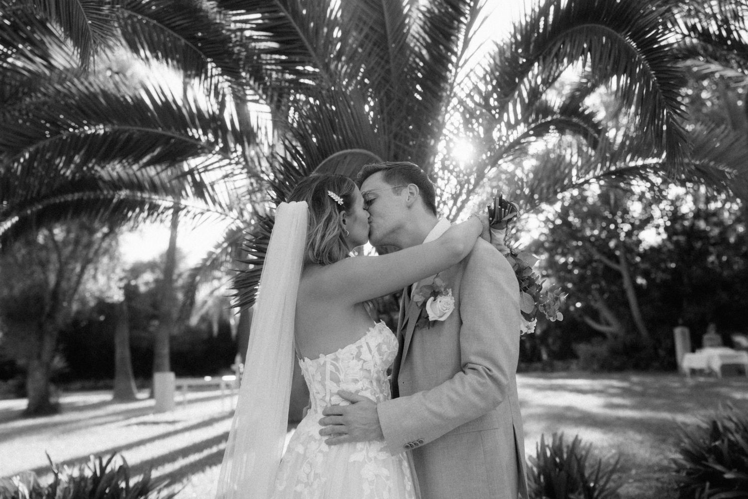 Black and white photo of bride and groom sharing a kiss under palm trees at their wedding.