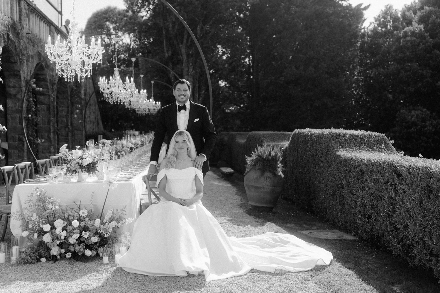 Black and white portrait of bride and groom posing beside outdoor wedding reception table with chandelier.