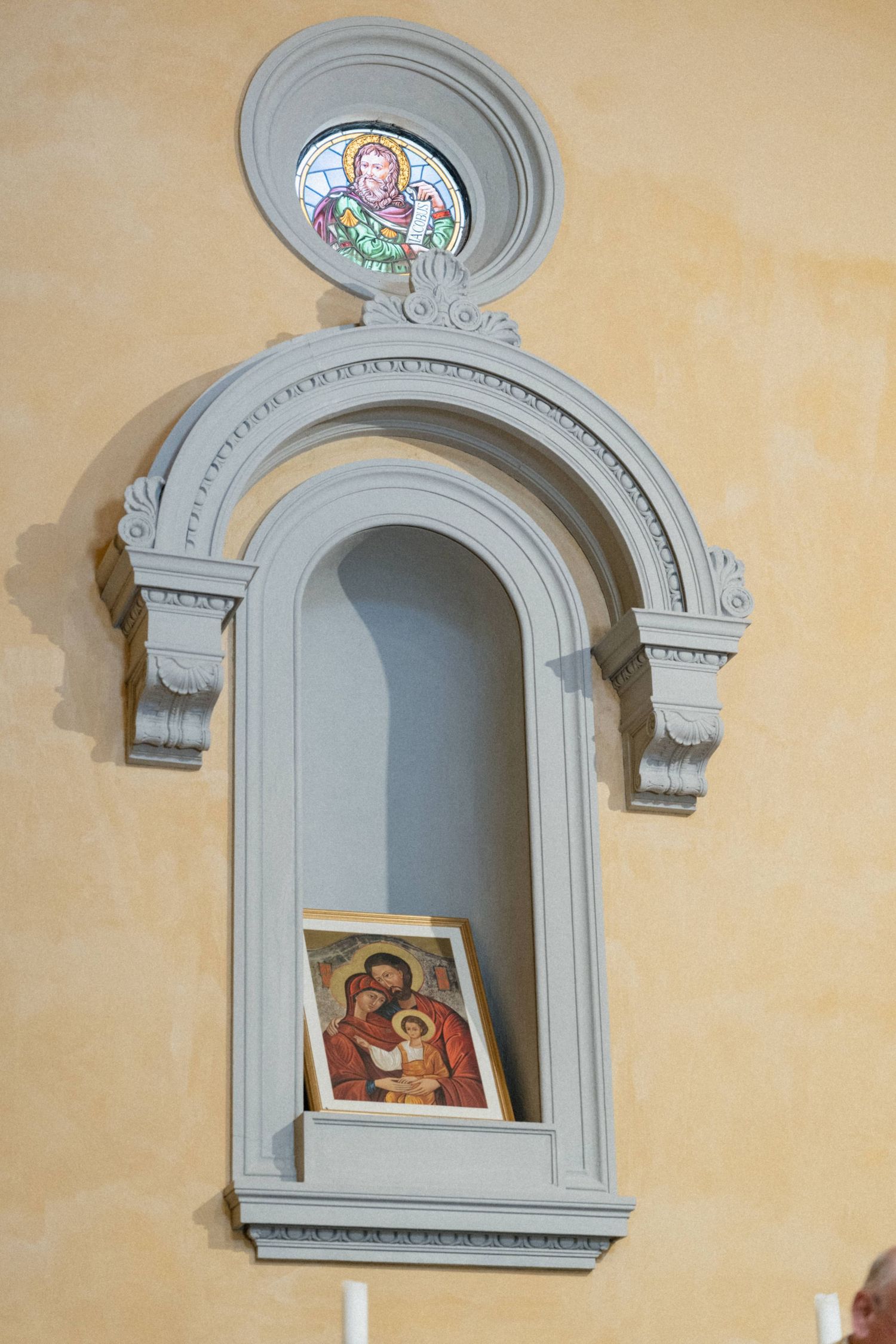 Religious icon displayed in ornate arched niche with stained glass angel above in church.