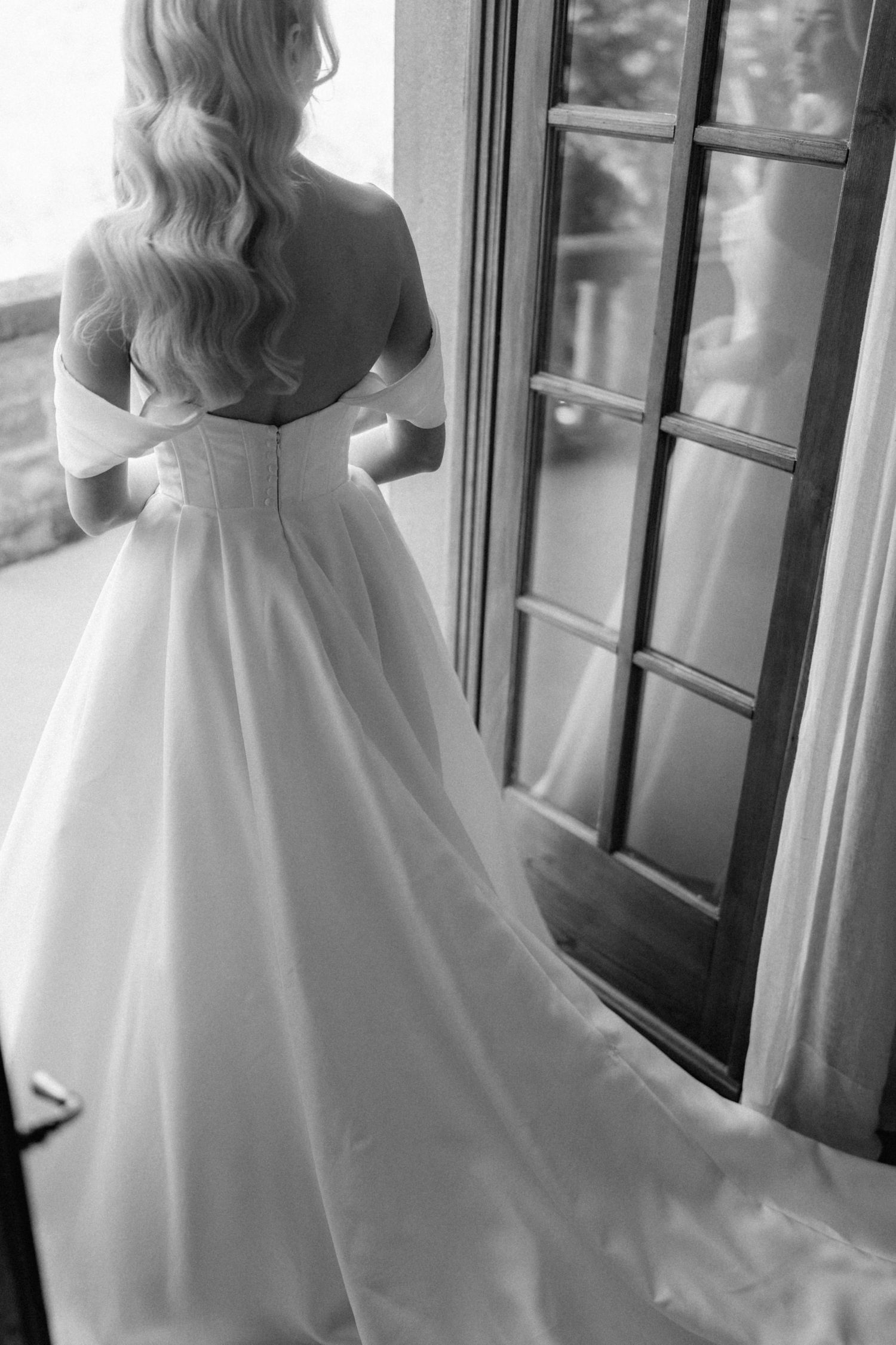 Black and white rear view of bride in off-shoulder ball gown standing by French doors.