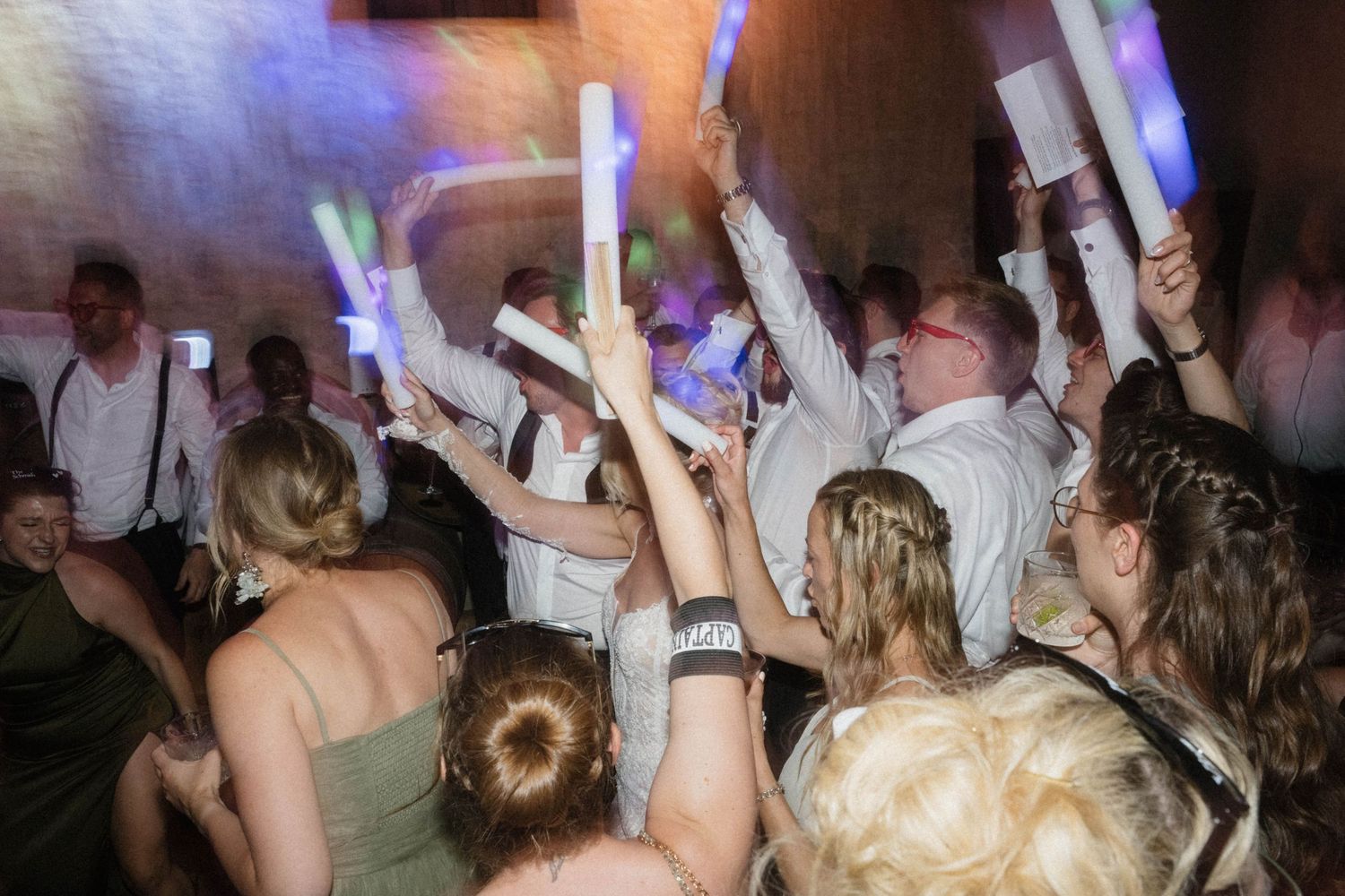 Wedding guests dancing and waving colorful glow sticks on crowded dance floor at reception party.