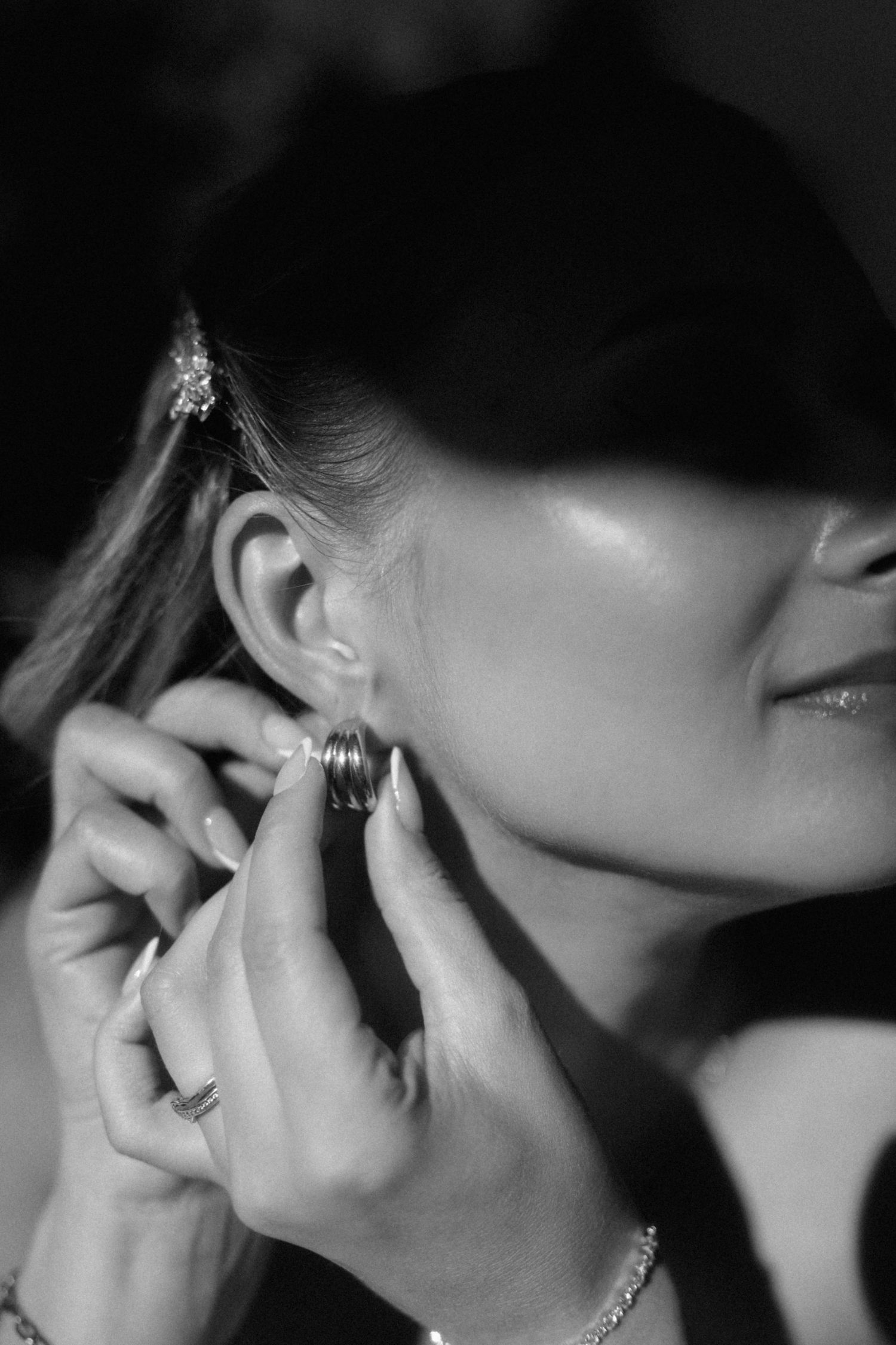 Black and white close-up of bride putting on gold hoop earring with bracelet on wrist.
