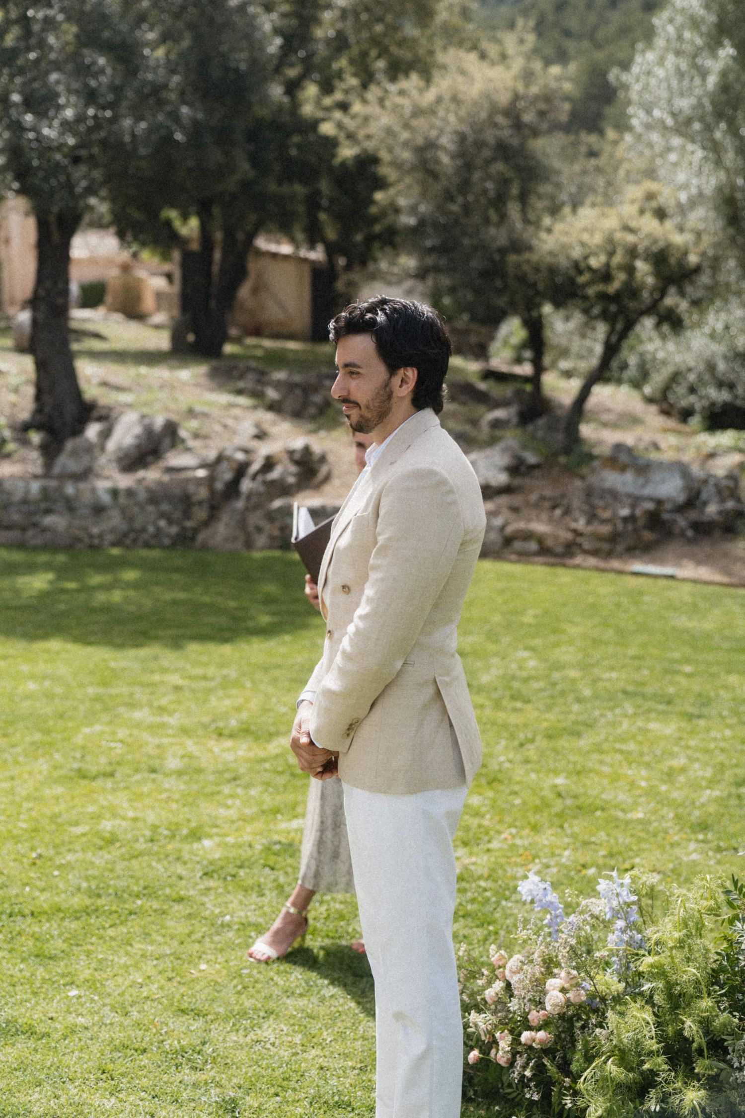 Handsome groom in beige linen blazer and white trousers stands on green lawn at outdoor garden wedding ceremony.