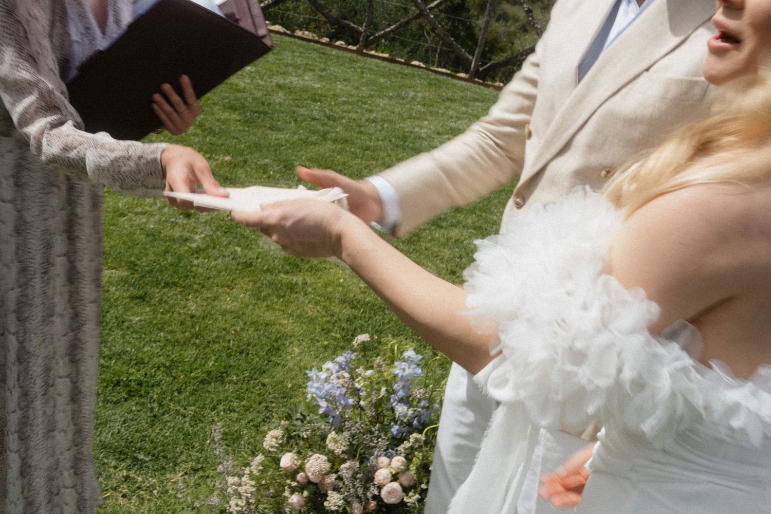 Bride in a white feathered dress reading vows during an outdoor garden wedding ceremony with a officiant.