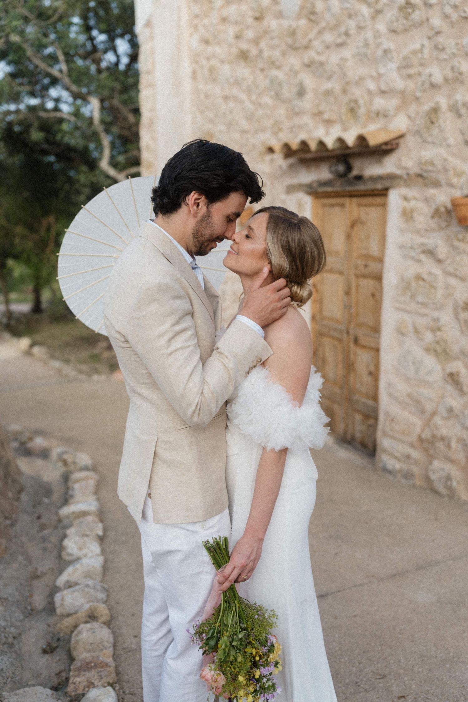 Bride and groom sharing romantic moment outside rustic stone building with wildflower bouquet.