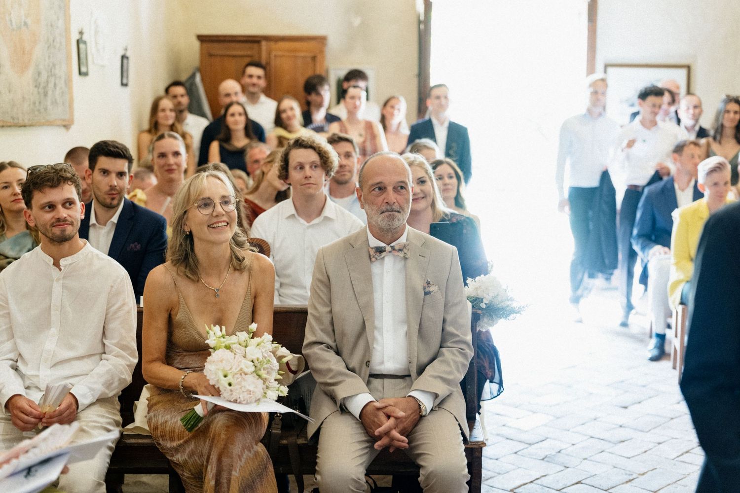 Wedding guests seated in a sunlit venue, smiling couple in foreground, bride holding white bouquet.