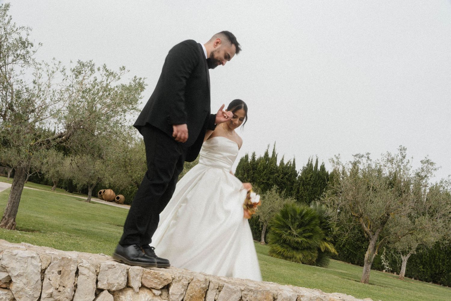 Bride and groom share a romantic moment outdoors, standing on a stone wall surrounded by lush greenery on their wedding day.