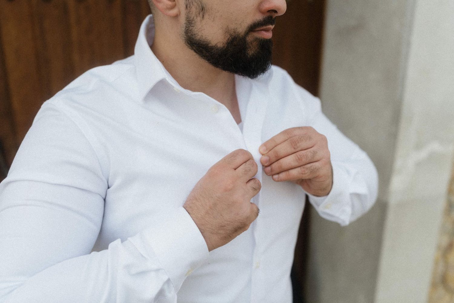 Bearded man in white dress shirt buttoning up collar, getting dressed for formal occasion.