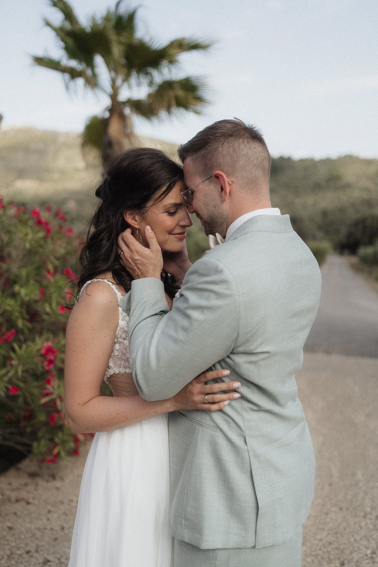 Bride and groom share a romantic embrace outdoors, with palm trees and red flowers in the background.