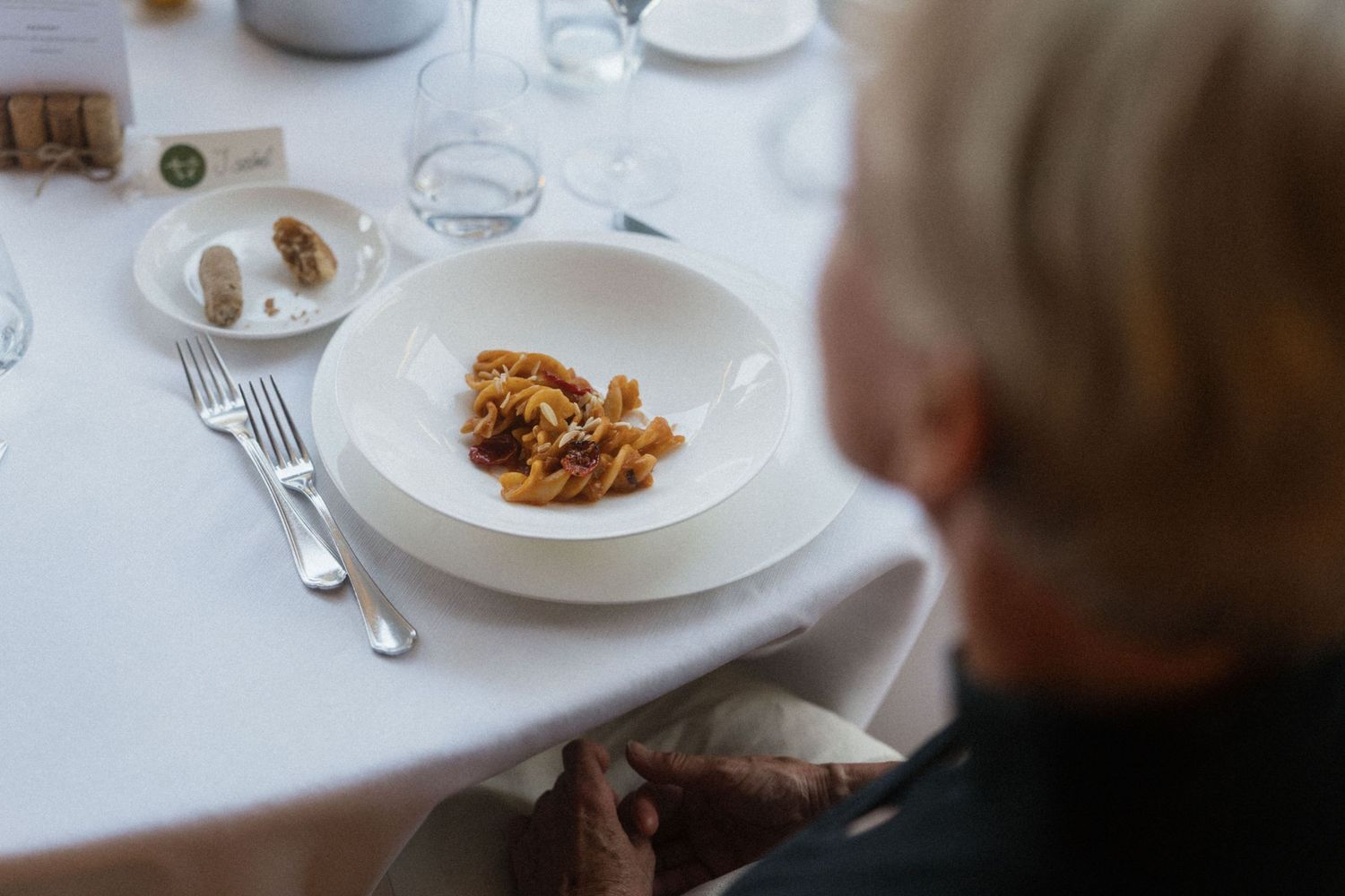 Elderly person dining at elegant table with white plate of pasta dish, fine restaurant setting with glassware.