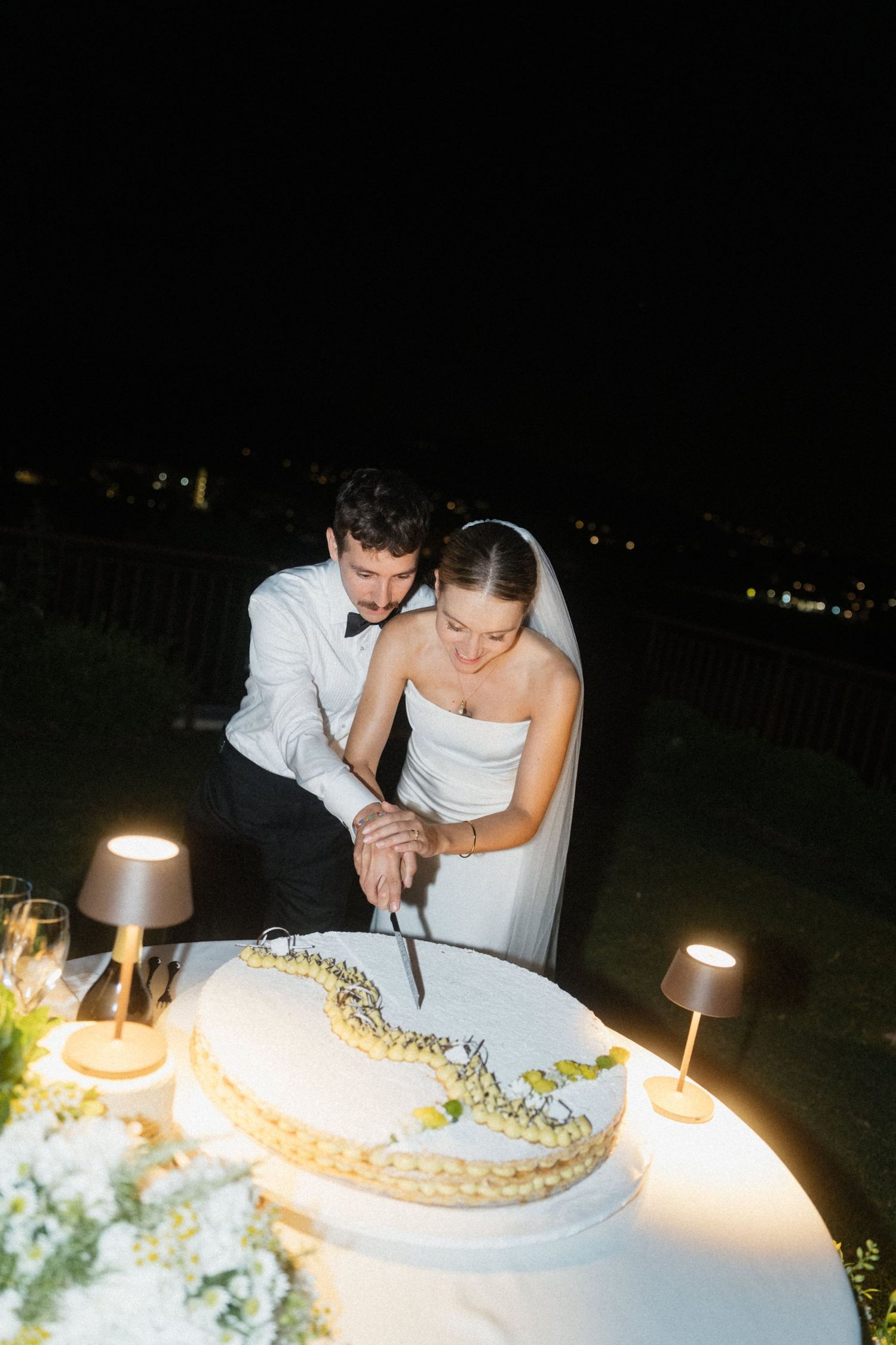 Bride and groom cutting their wedding cake together at night, surrounded by soft glowing table lamps.