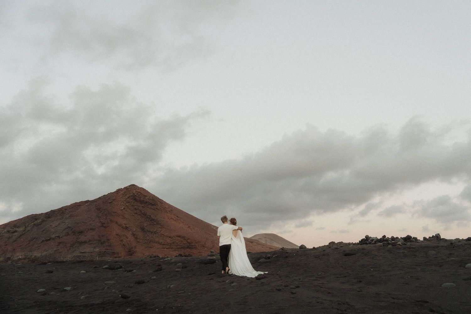 Wedding couple embracing on dark volcanic landscape with red mountain and cloudy sky in background.