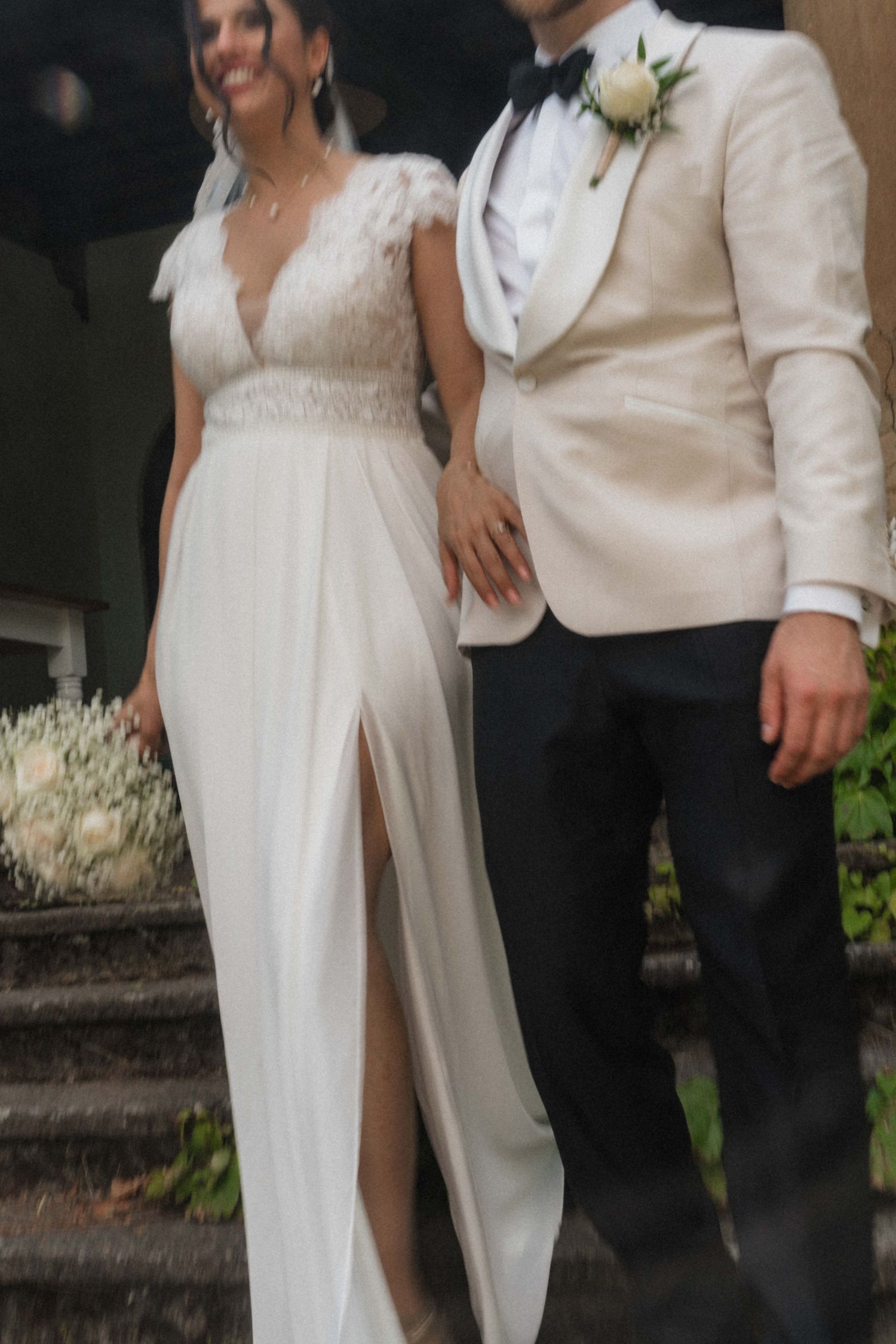 Bride and groom standing together, bride in lace wedding gown with slit, groom in ivory tuxedo with boutonniere.