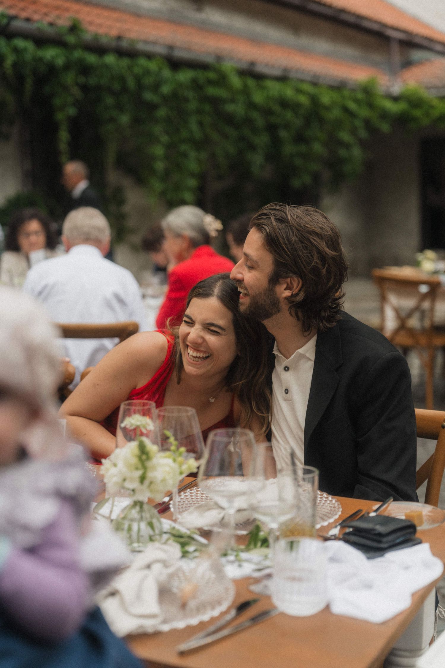 Laughing couple embracing at outdoor wedding reception dinner table surrounded by guests.