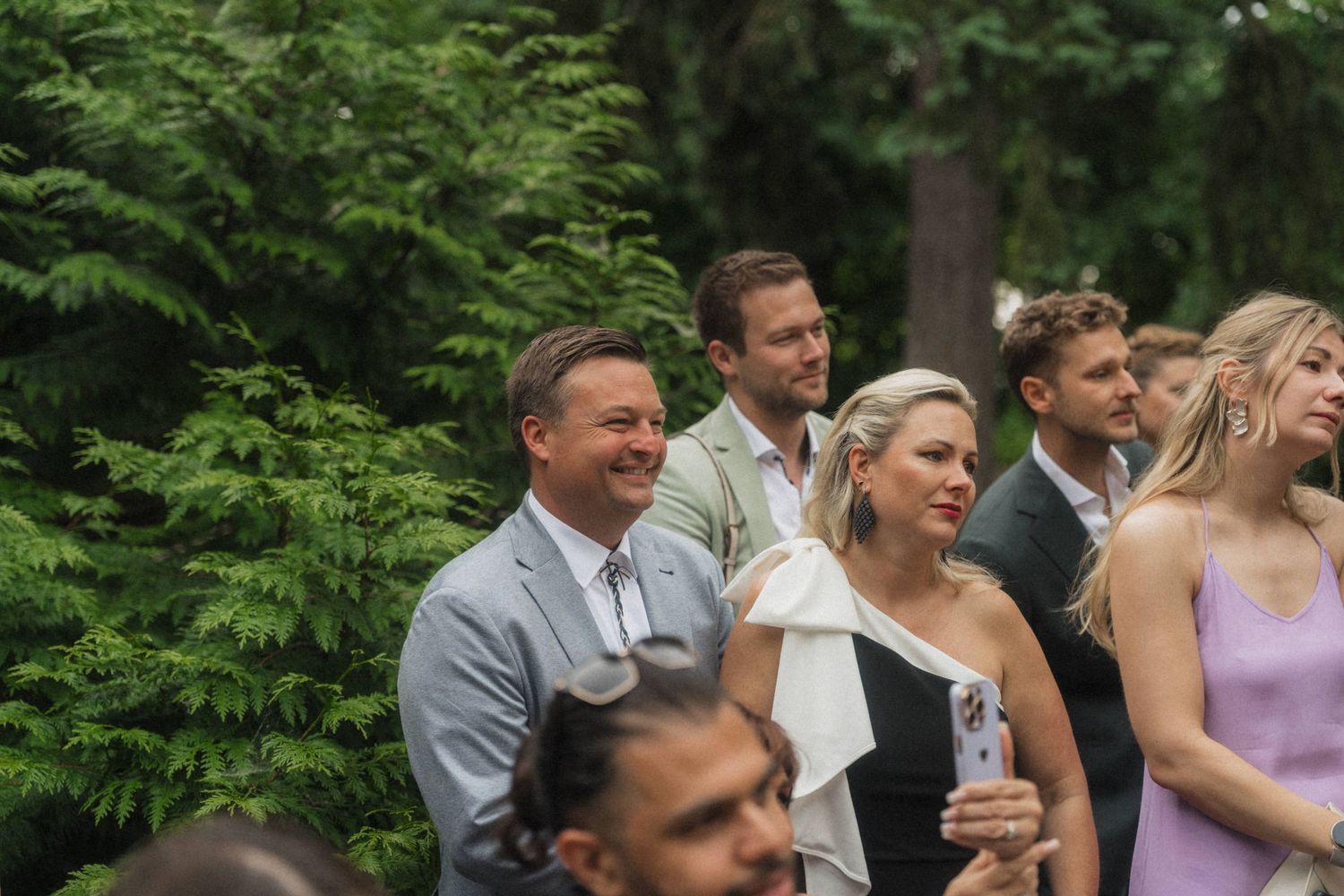 Smiling wedding guests in elegant attire watching outdoor ceremony surrounded by tall green trees.