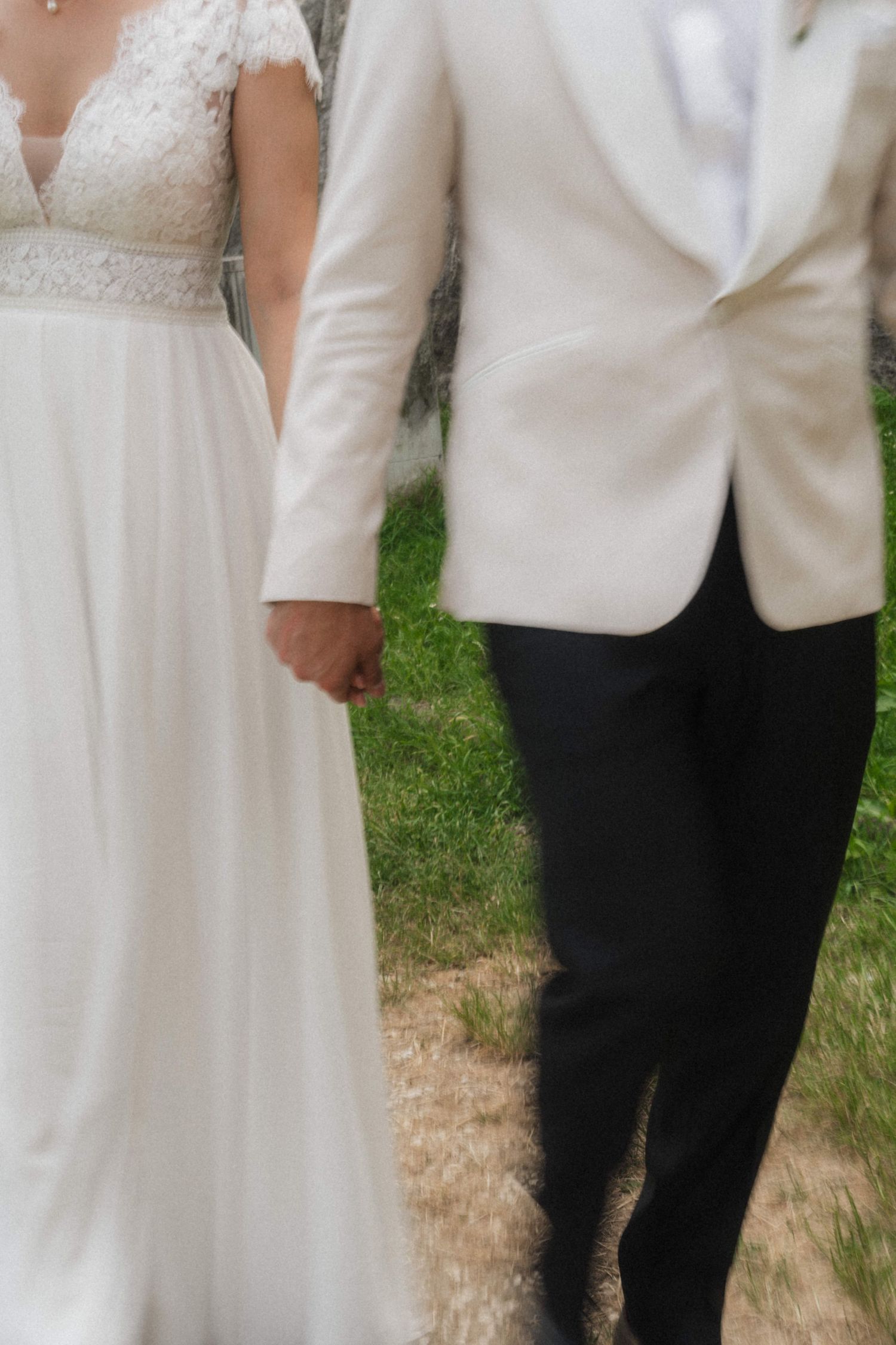 Bride and groom holding hands in wedding attire, white lace dress and ivory tuxedo jacket, walking outdoors.